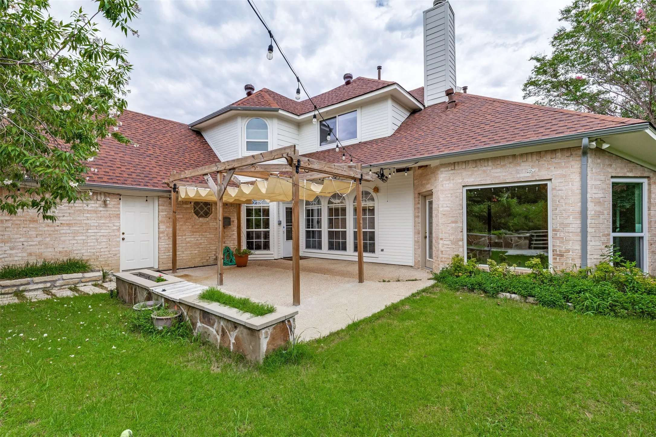 Expansive backyard featuring a stone patio with a wood pergola, red shingle roofing, light-colored brick exterior, and a built-in stone planter