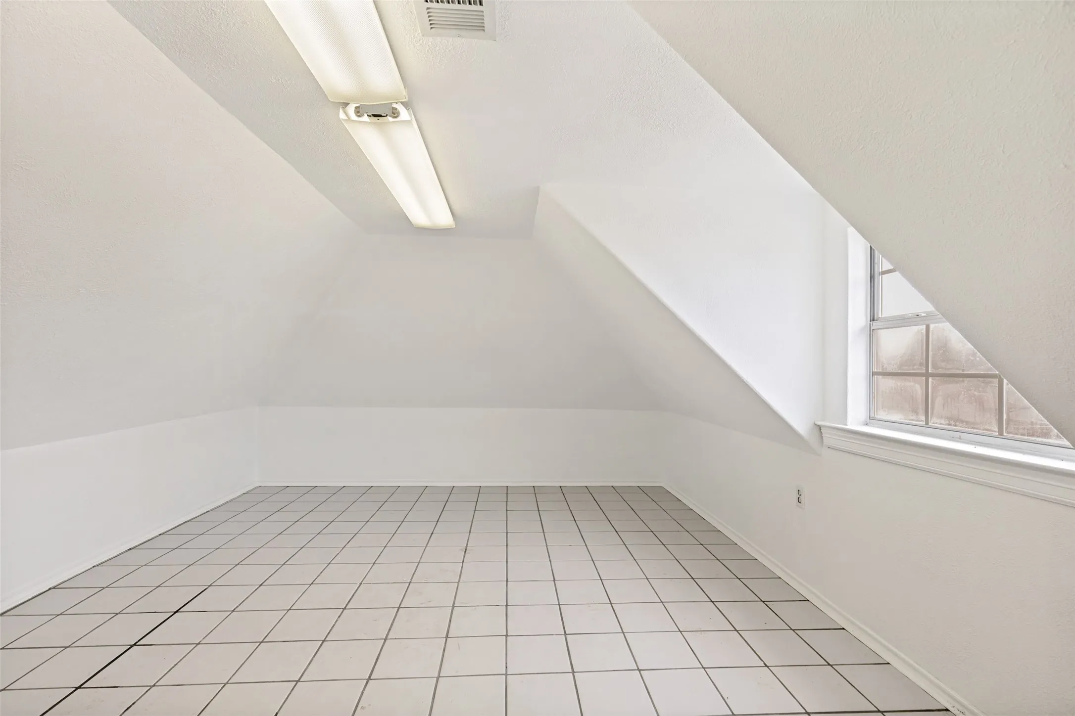 Attic space featuring sloped ceilings, white square tile flooring, and a single window with privacy glass blocks