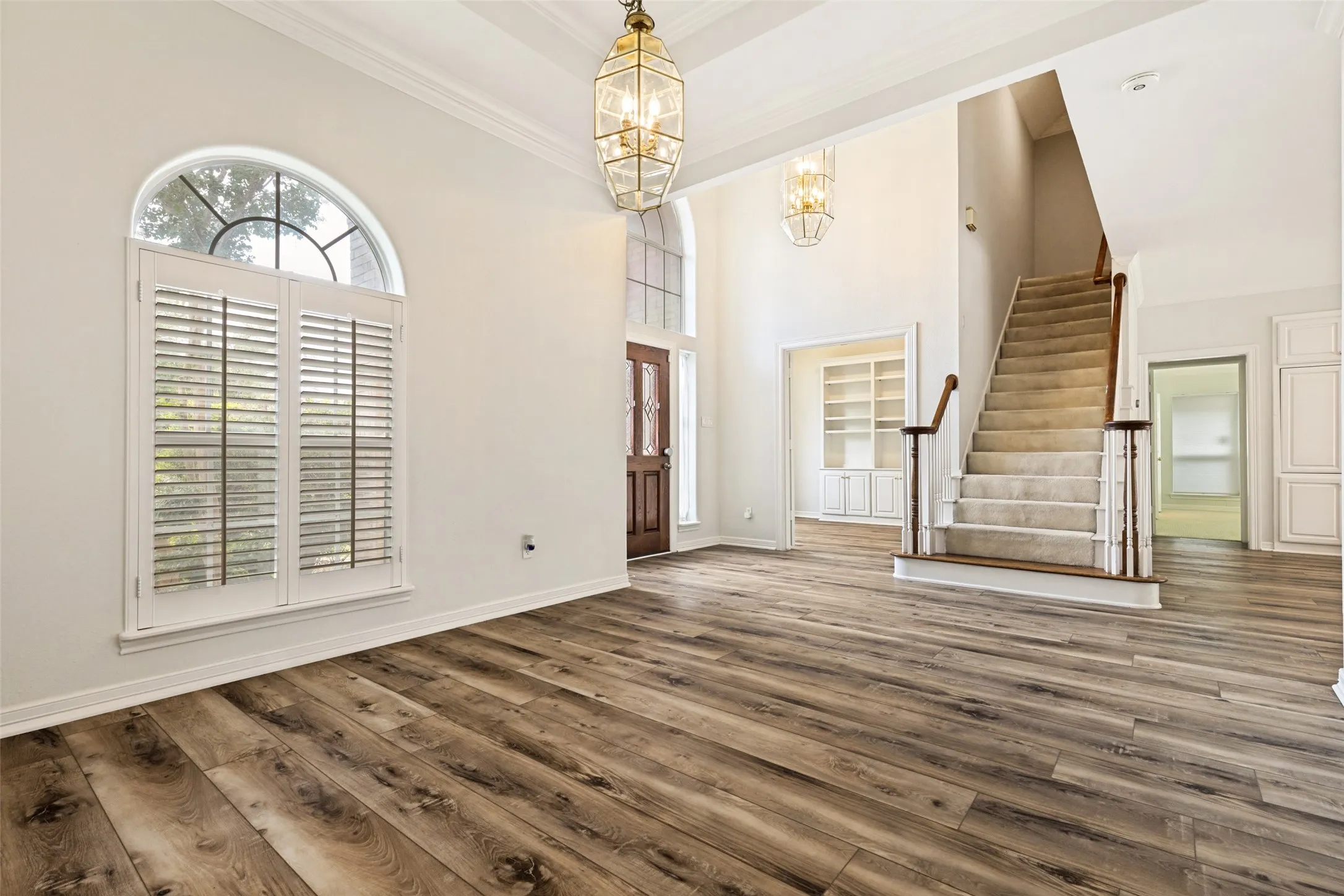 Foyer featuring wood-finish flooring, a grand staircase with wood railing, custom plantation shutters, two-story ceilings with decorative light fixtures, and a front entry door with a transom window