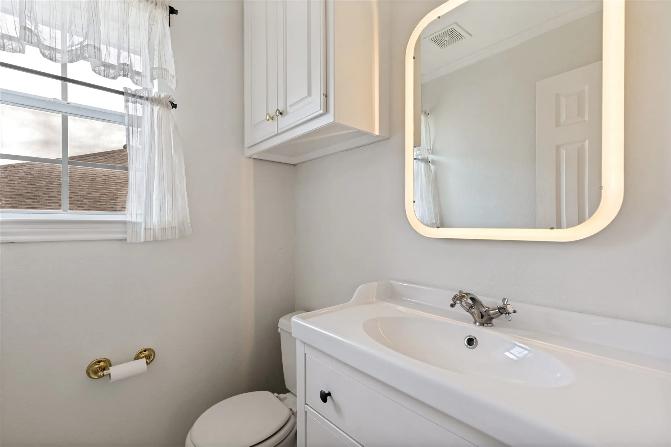 Bathroom featuring a white vanity with integrated sink, chrome faucet, illuminated mirror, and overhead cabinetry