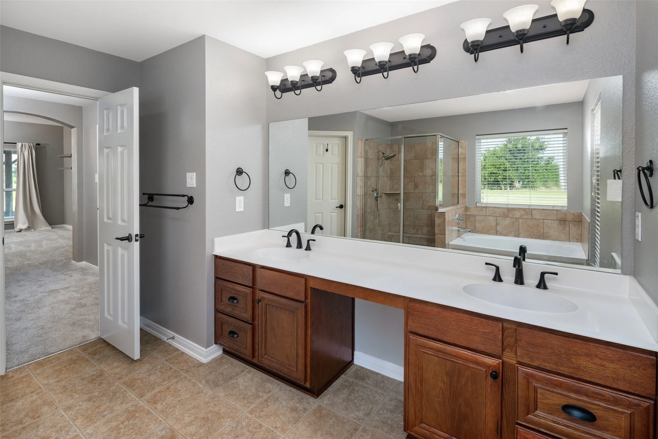 Bathroom featuring a dual vanity with wood cabinetry and white countertops, a tiled walk-in shower with a glass enclosure, a built-in tub with tile surround, and a broad window