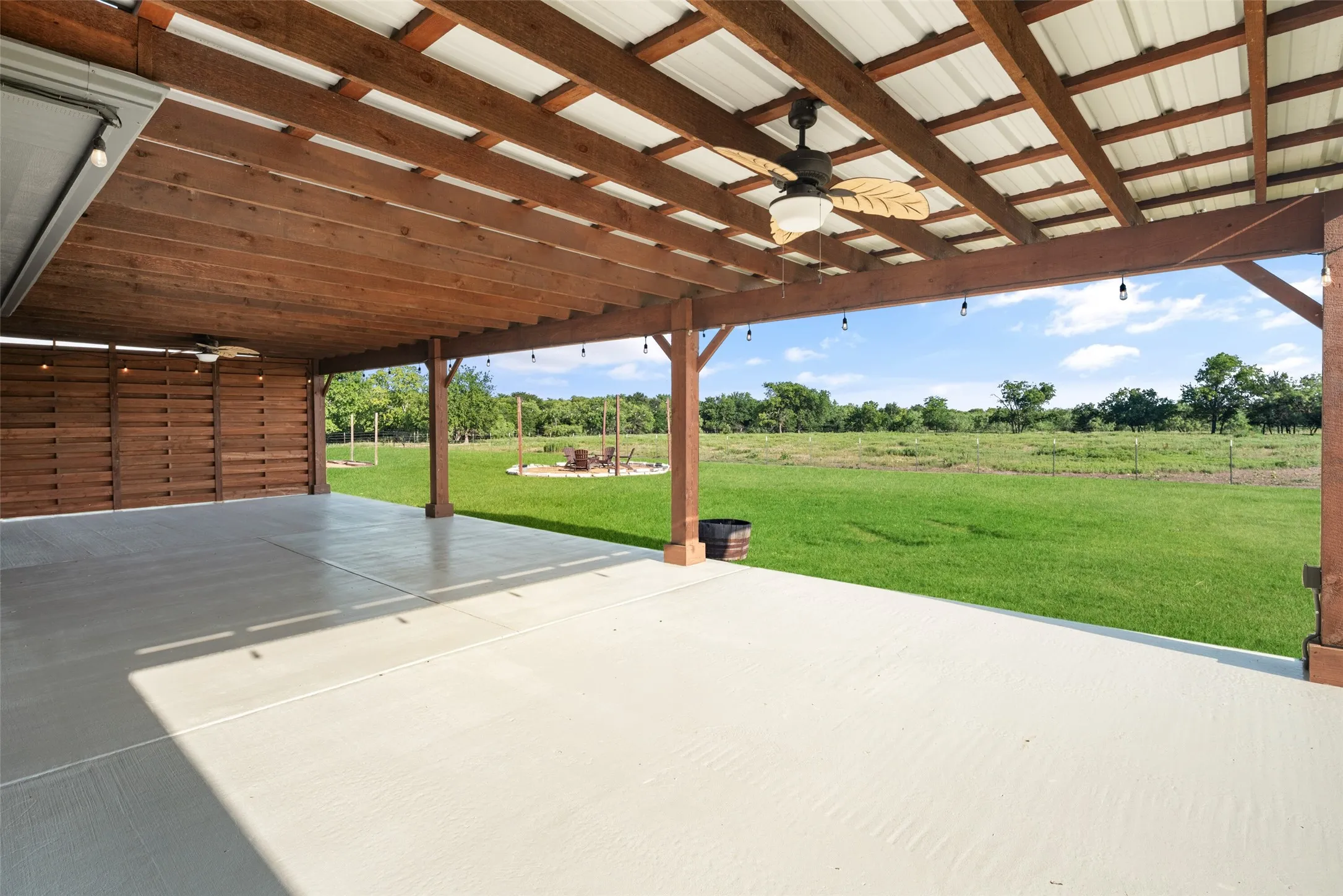Expansive covered patio featuring a wood-beam ceiling with corrugated metal roofing, an integrated ceiling fan, and string lighting