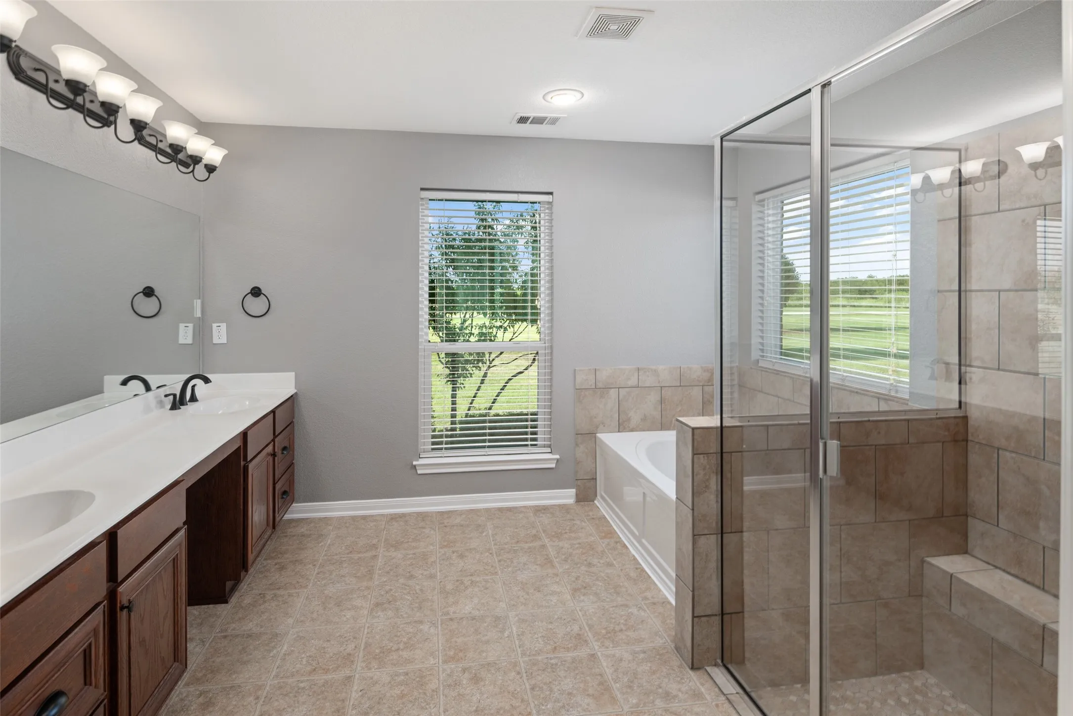 Spacious bathroom featuring a dual vanity with dark wood cabinetry, a large built-in mirror, a glass-enclosed shower with tiled walls and a built-in bench, a separate bathtub, and tile flooring