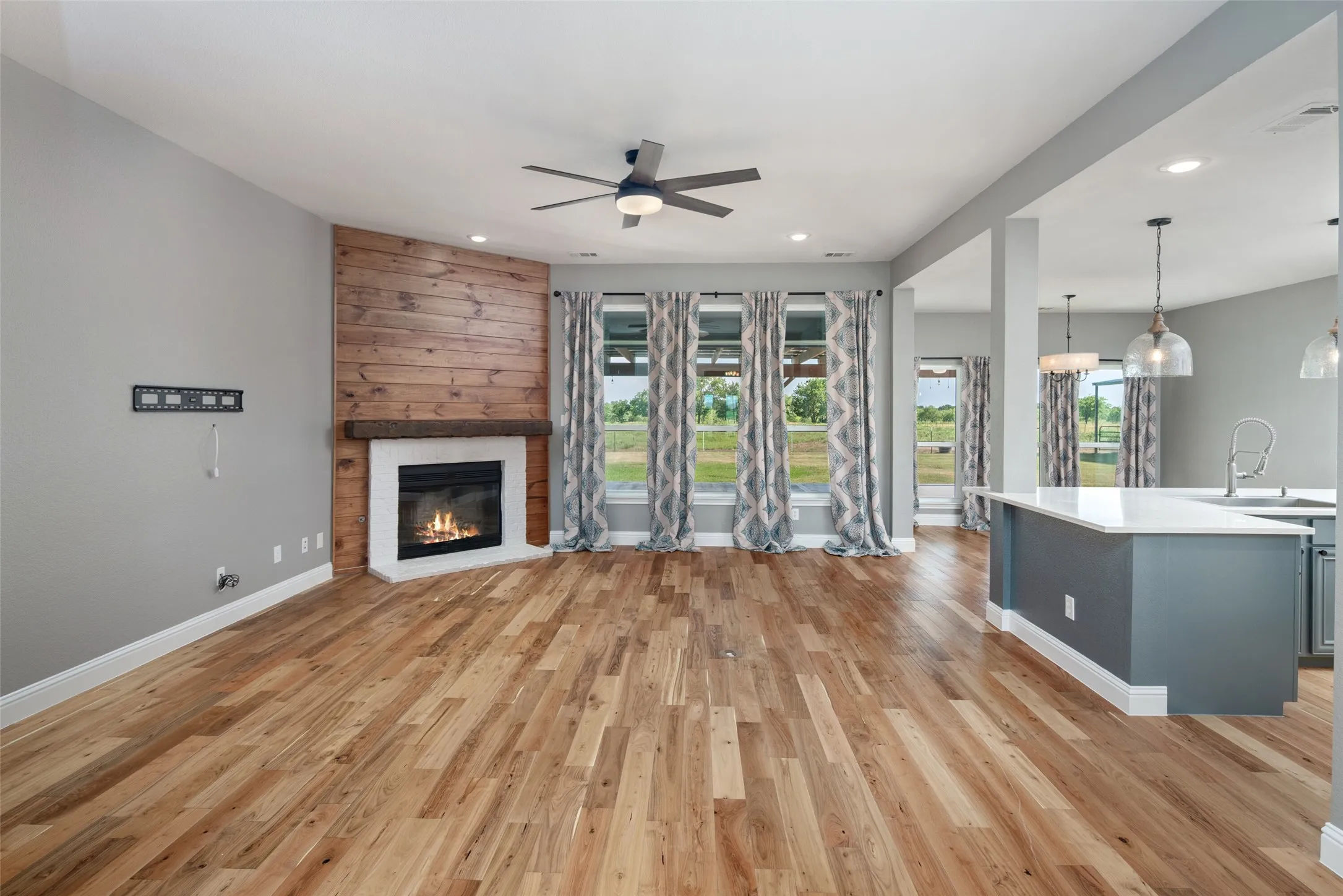Spacious living area featuring wood-finish flooring, a corner fireplace with a rustic wood-plank accent wall and white mantel, and a ceiling fan