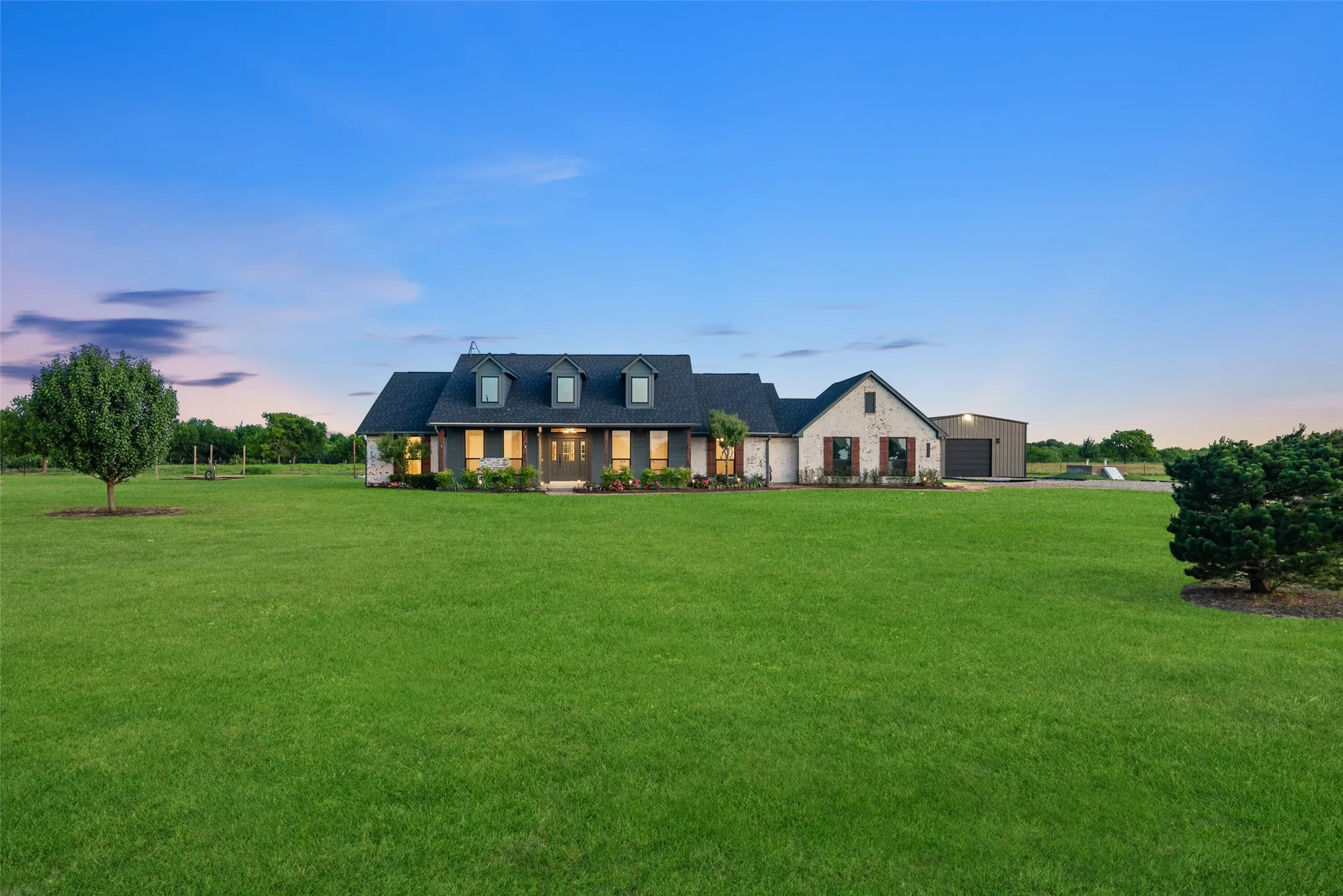 Expansive green lawn surrounds this property featuring a main residence with a dark shingle roof, multiple dormer windows, and a stone facade accent