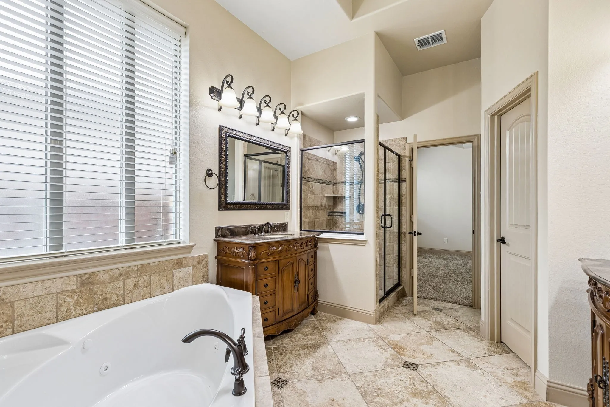Spacious primary bathroom featuring a white soaking tub with oil-rubbed bronze fixtures, a separate shower with a glass enclosure and tiled walls, a wood vanity with an integrated sink and dark countertop, and decorative floor tiling