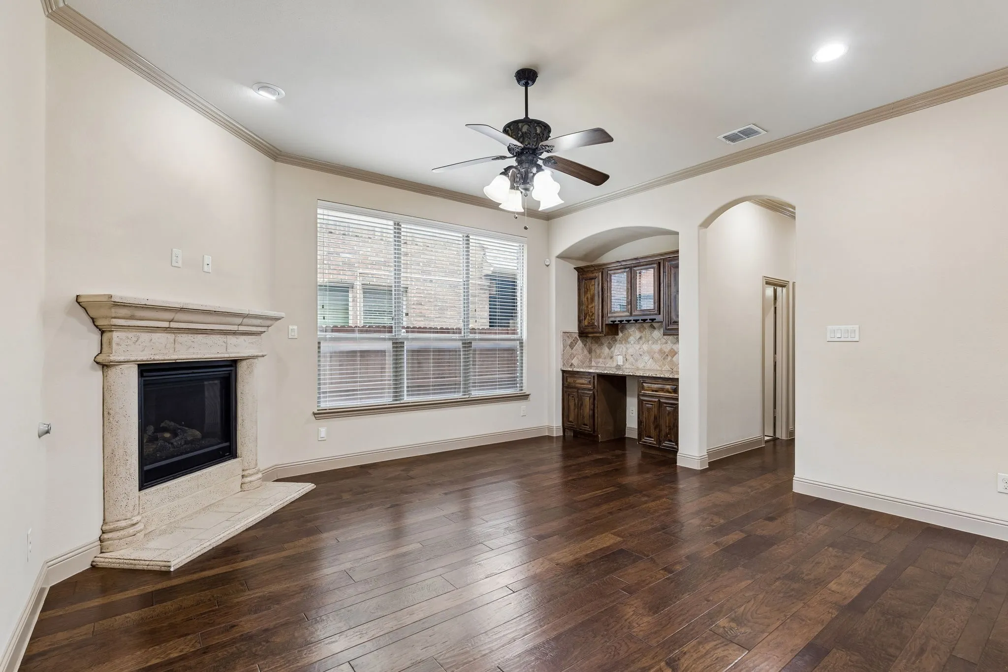 Hardwood-style flooring throughout this room, featuring a stone-clad fireplace with mantel, crown molding, and a built-in wet bar with dark wood cabinetry and a tiled backsplash