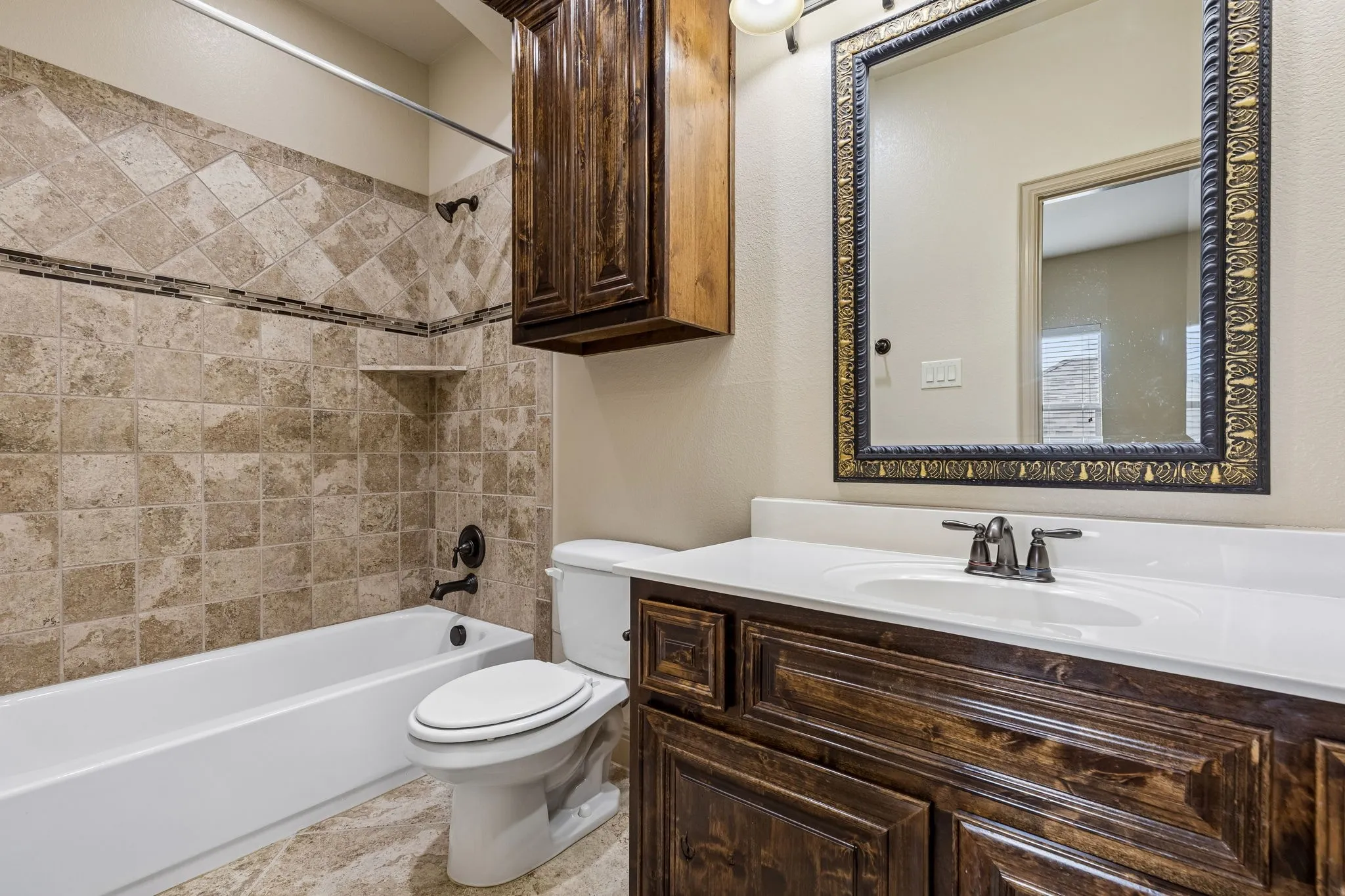 Ensuite bathroom featuring a white bathtub, tiled shower surround with accent band, dark wood vanity with white countertop and integrated sink, and an ornate framed mirror