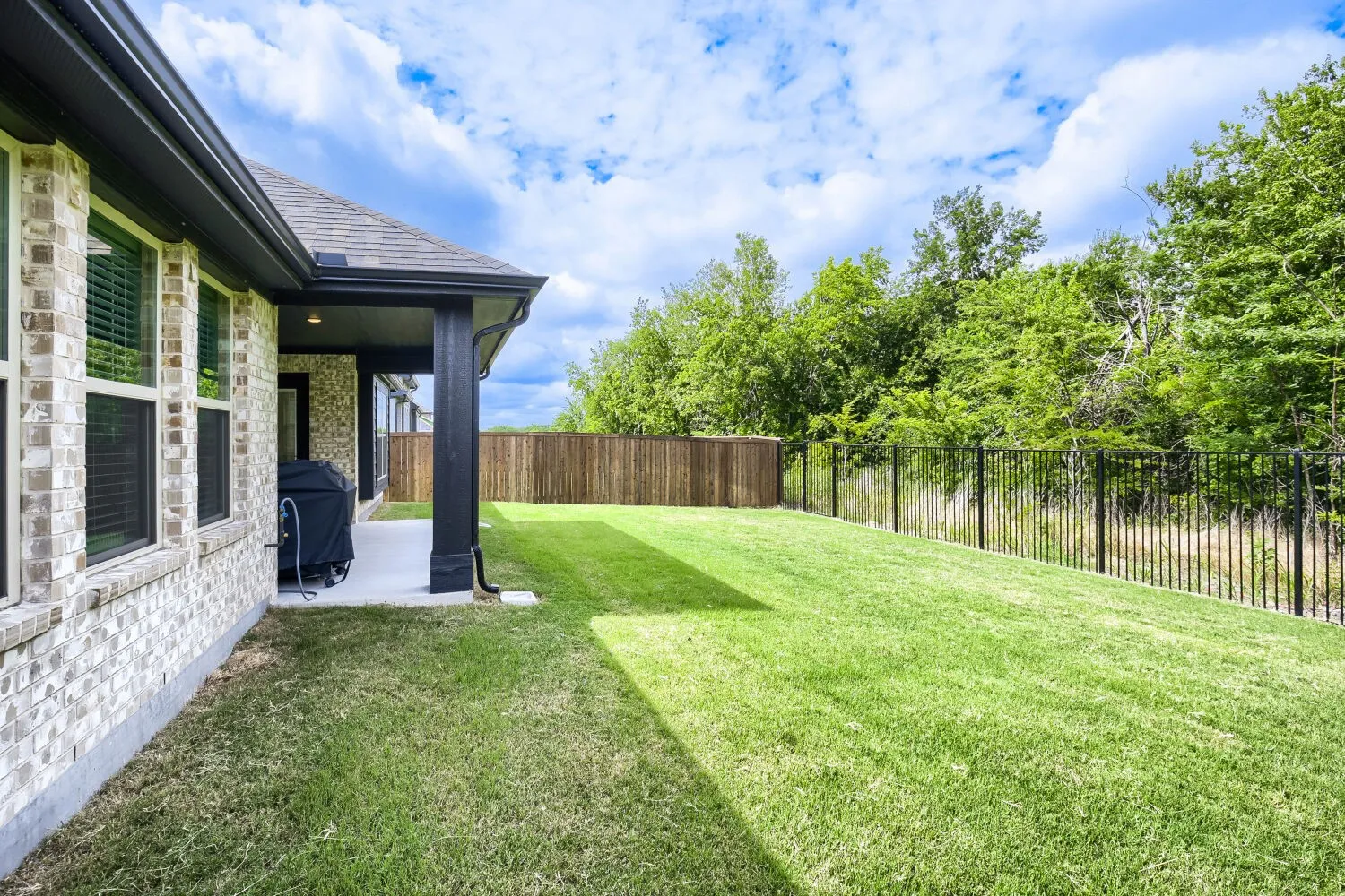 Expansive backyard featuring a manicured lawn, covered patio, two fencing types, and mature trees beyond the property line