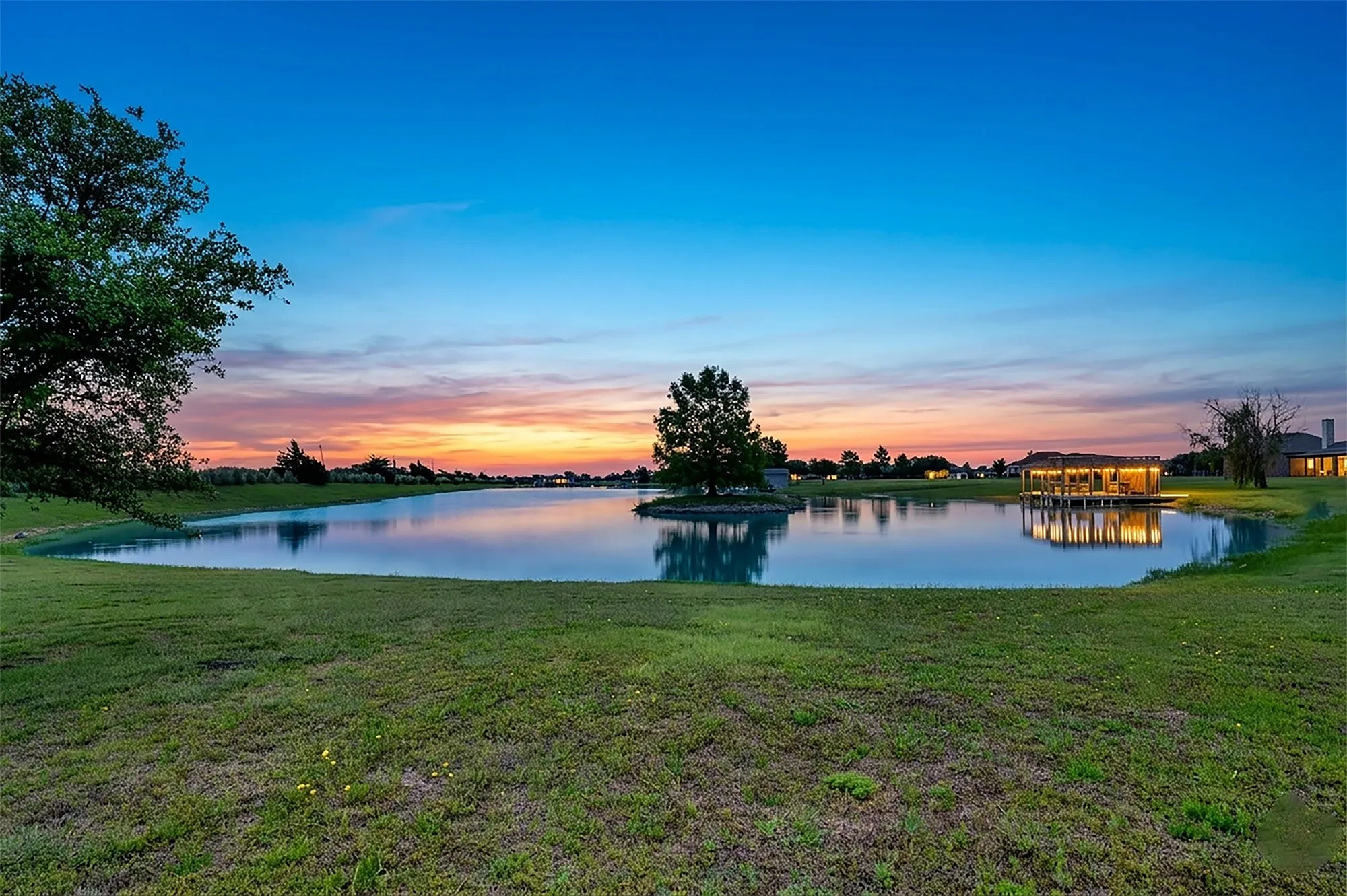 Expansive lawn bordering a serene body of water, featuring a private dock structure with overhead lighting and a central island with mature trees