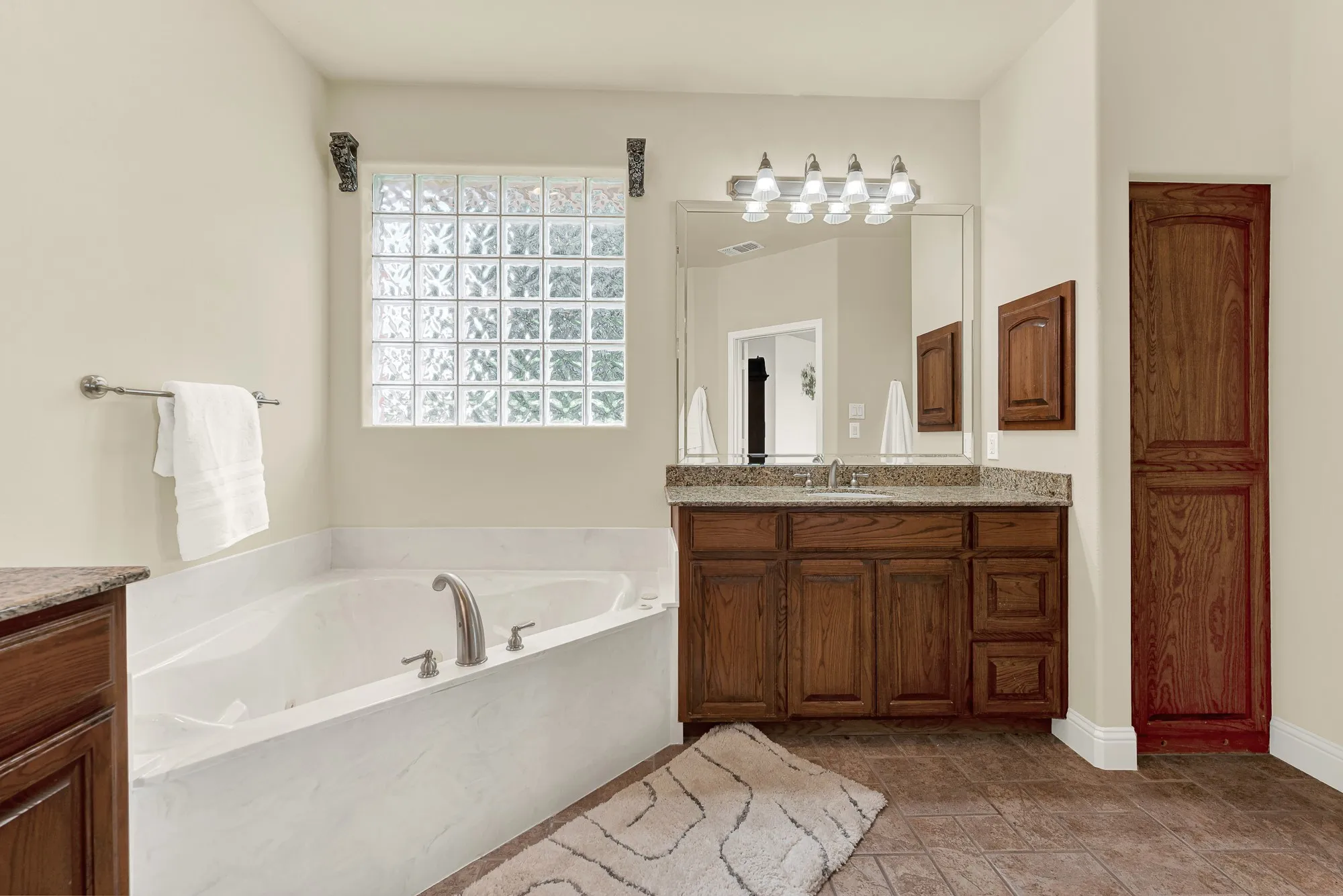 Spacious bathroom featuring a built-in soaking tub, a glass block window, dark wood vanity with a stone countertop, a large mirror, and ample built-in storage cabinetry