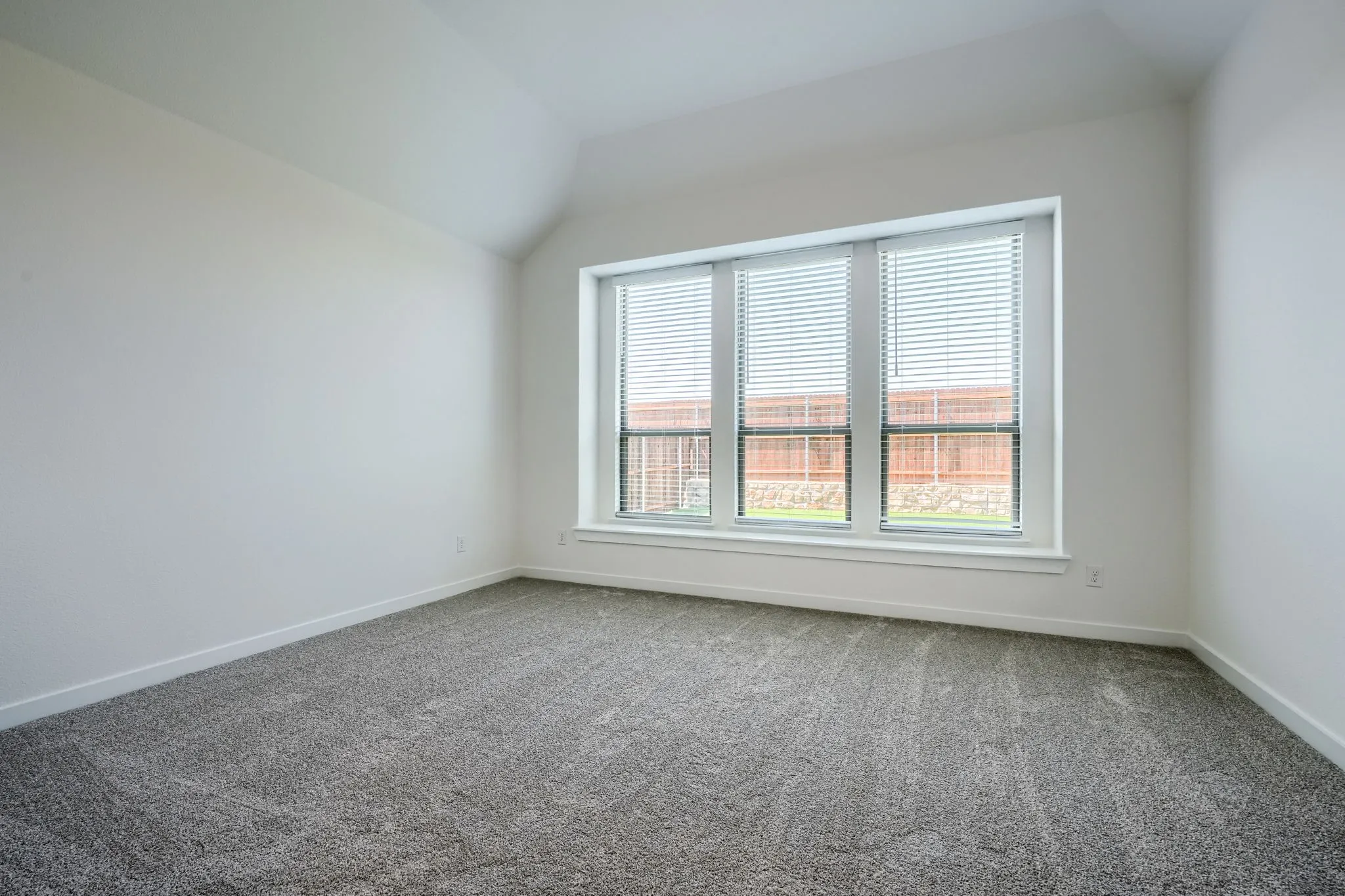 Room featuring a large triple window with blinds, vaulted ceiling, neutral wall paint, white baseboards, and patterned carpet flooring
