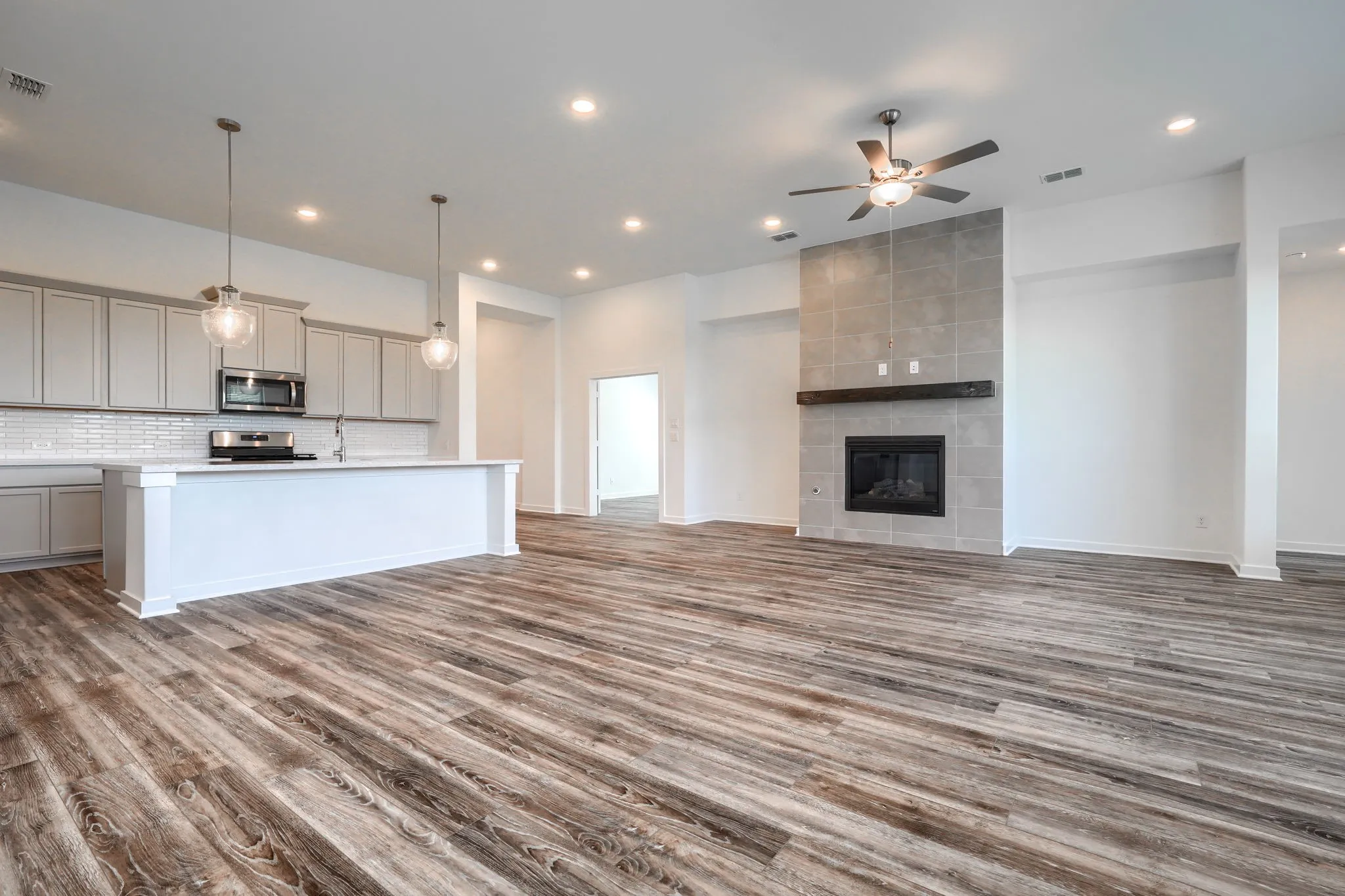 Open-concept living space featuring wood-finish flooring, a kitchen island with pendant lighting, recessed ceiling lights, a tiled fireplace with a wood mantel, and a ceiling fan