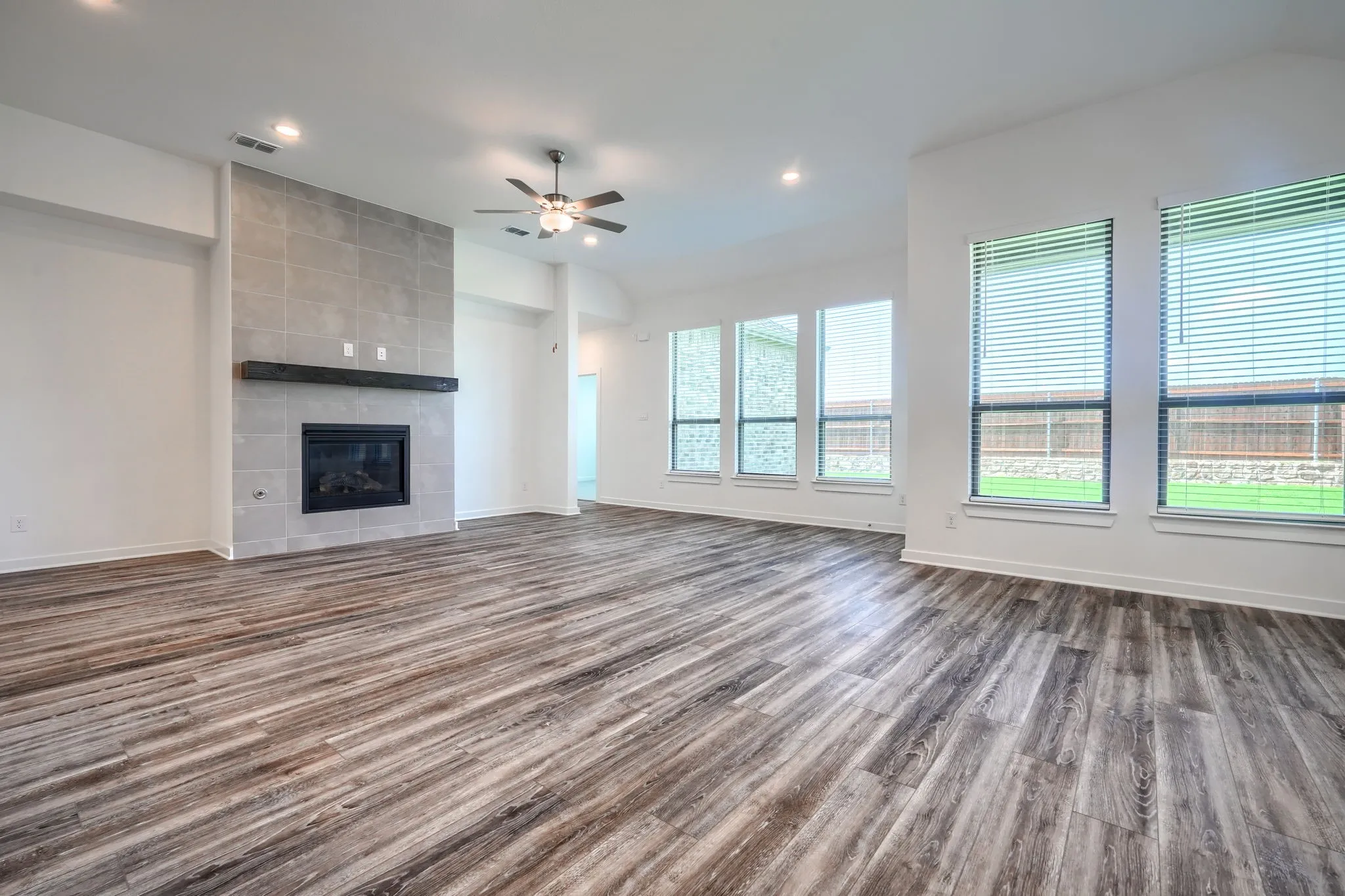 Spacious room featuring wood-finish flooring, a contemporary fireplace with a tiled surround and dark wood mantel, and multiple large windows with blinds