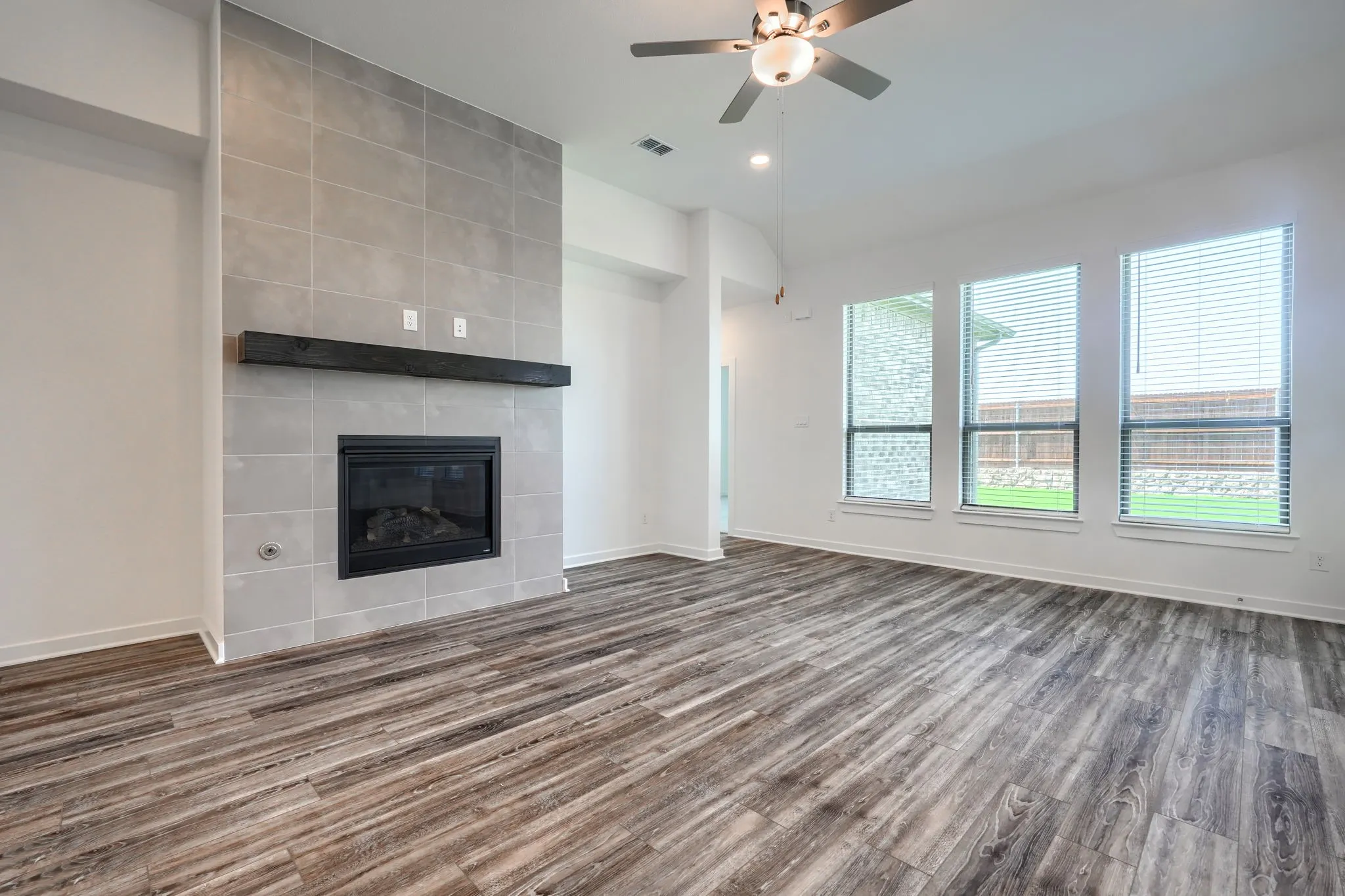 Living area featuring a floor-to-ceiling tiled fireplace with a dark wood mantel, wood-finish flooring, multiple windows with blinds, and a ceiling fan with integrated lighting