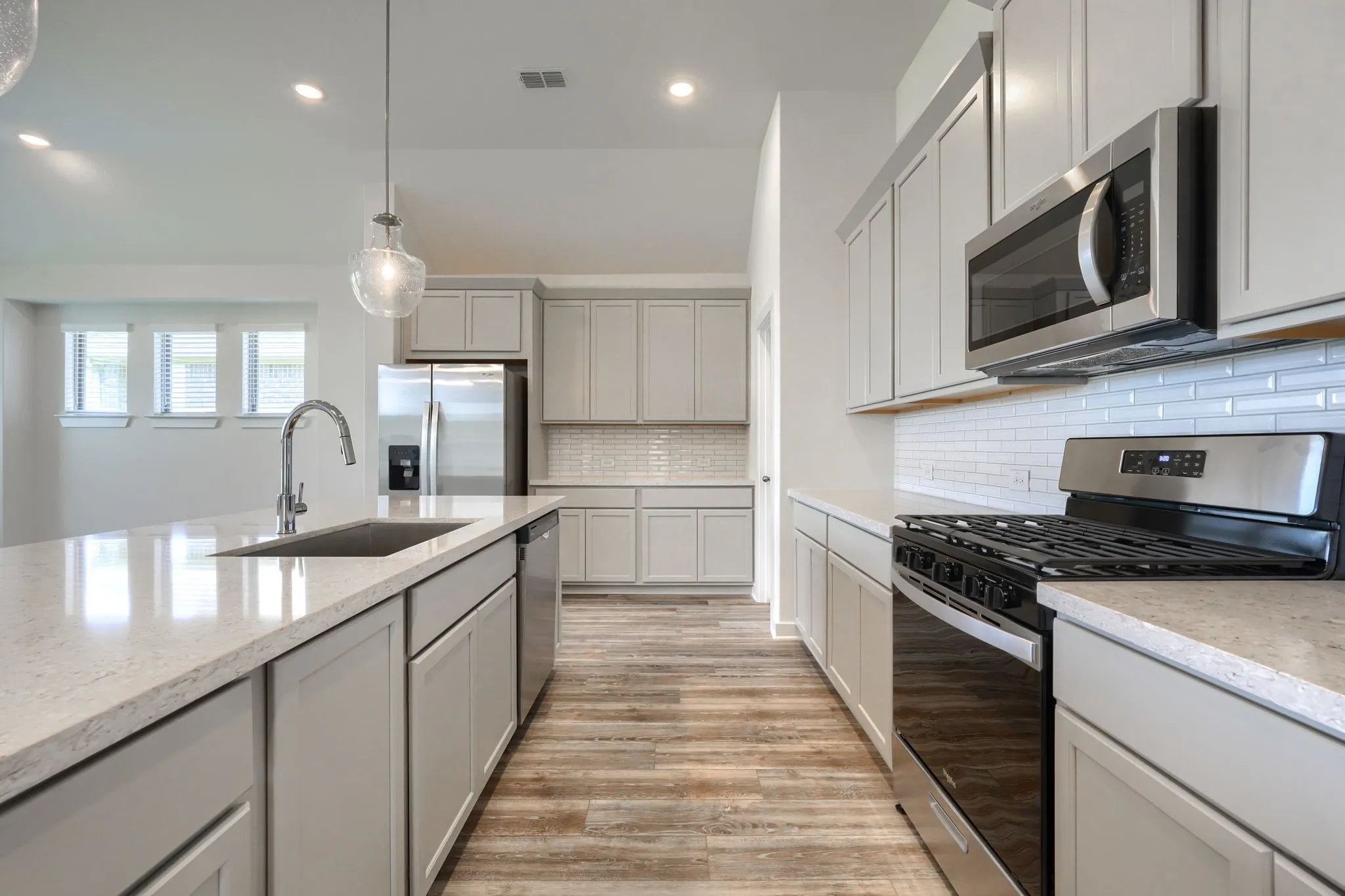 Kitchen featuring a central island with an undermount sink, stainless steel appliances, light gray cabinetry, wood-finish flooring, and a white subway tile backsplash
