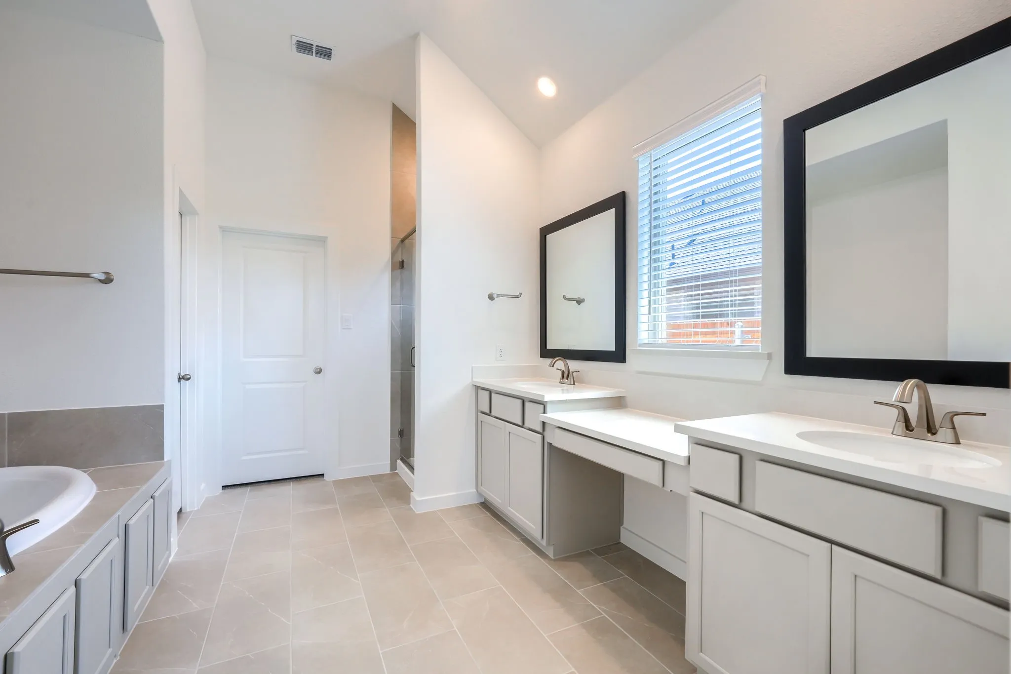 Spacious bathroom featuring light-toned tile flooring, dual vanities with integrated sinks and brush nickel fixtures, a built-in makeup vanity, a window with blinds, and a glass-enclosed shower