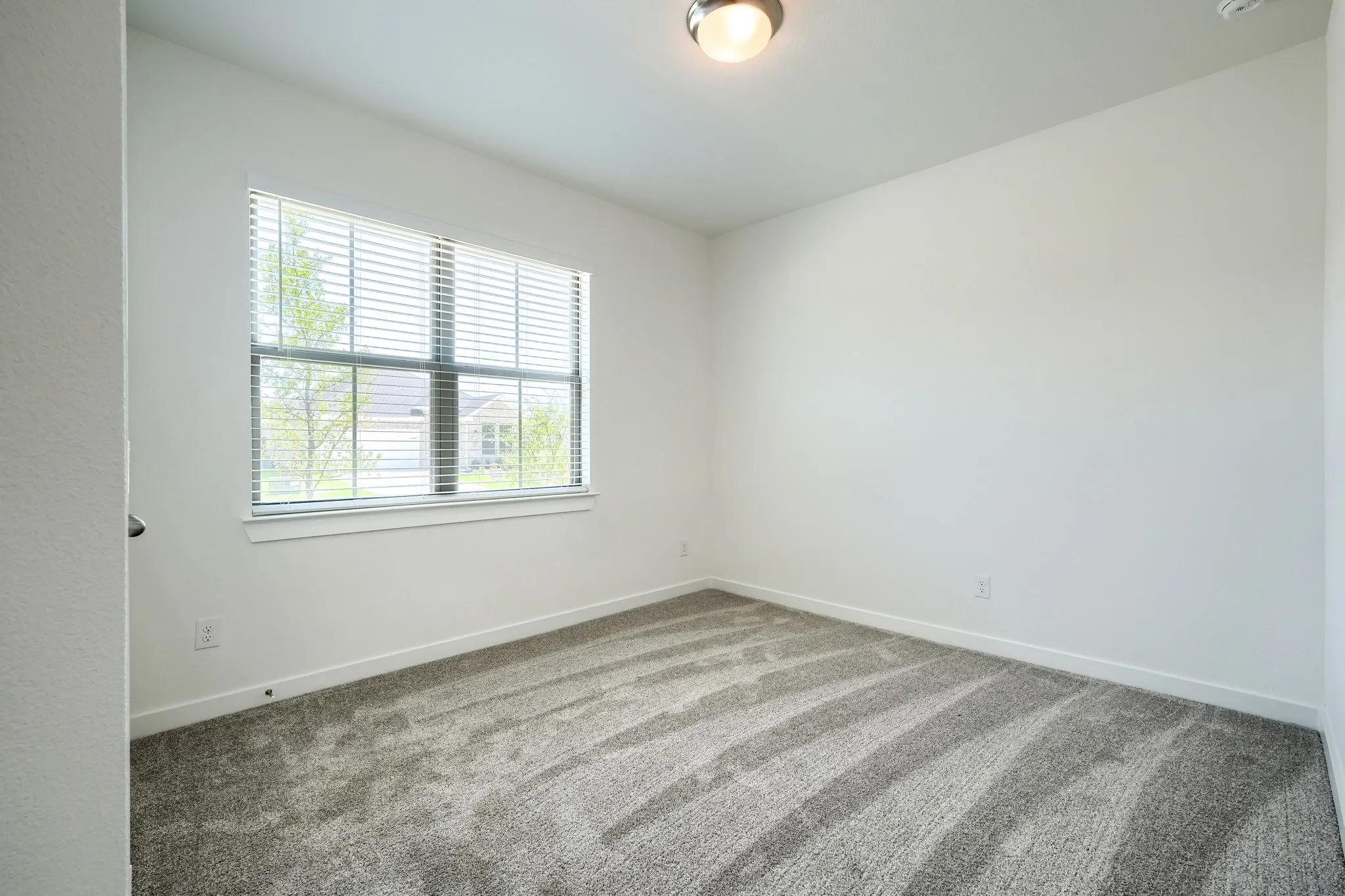 Bright interior space featuring a window with white blinds, grey patterned carpet, white walls, white trim, and a ceiling-mounted light fixture