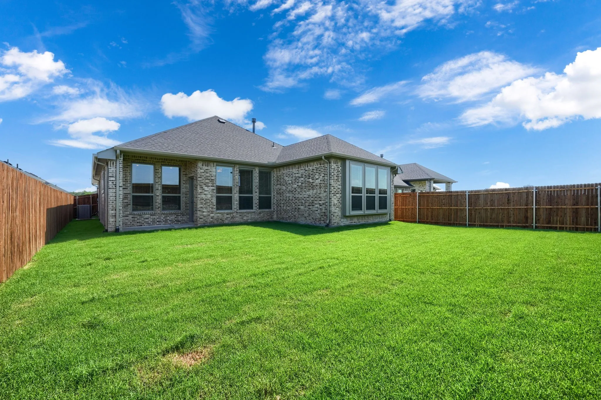 Expansive backyard featuring a manicured lawn, full privacy fencing, a brick exterior, a covered patio area, and multiple large windows
