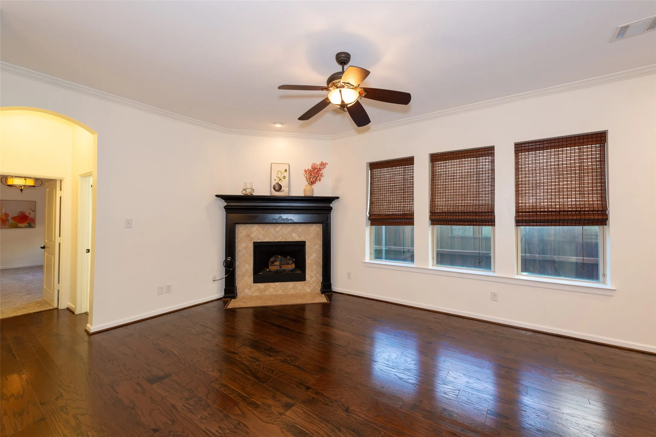 Living area featuring dark wood-finish flooring, a fireplace with a tiled surround and dark wood mantel, three windows with woven blinds, a ceiling fan with integrated lighting, and crown molding