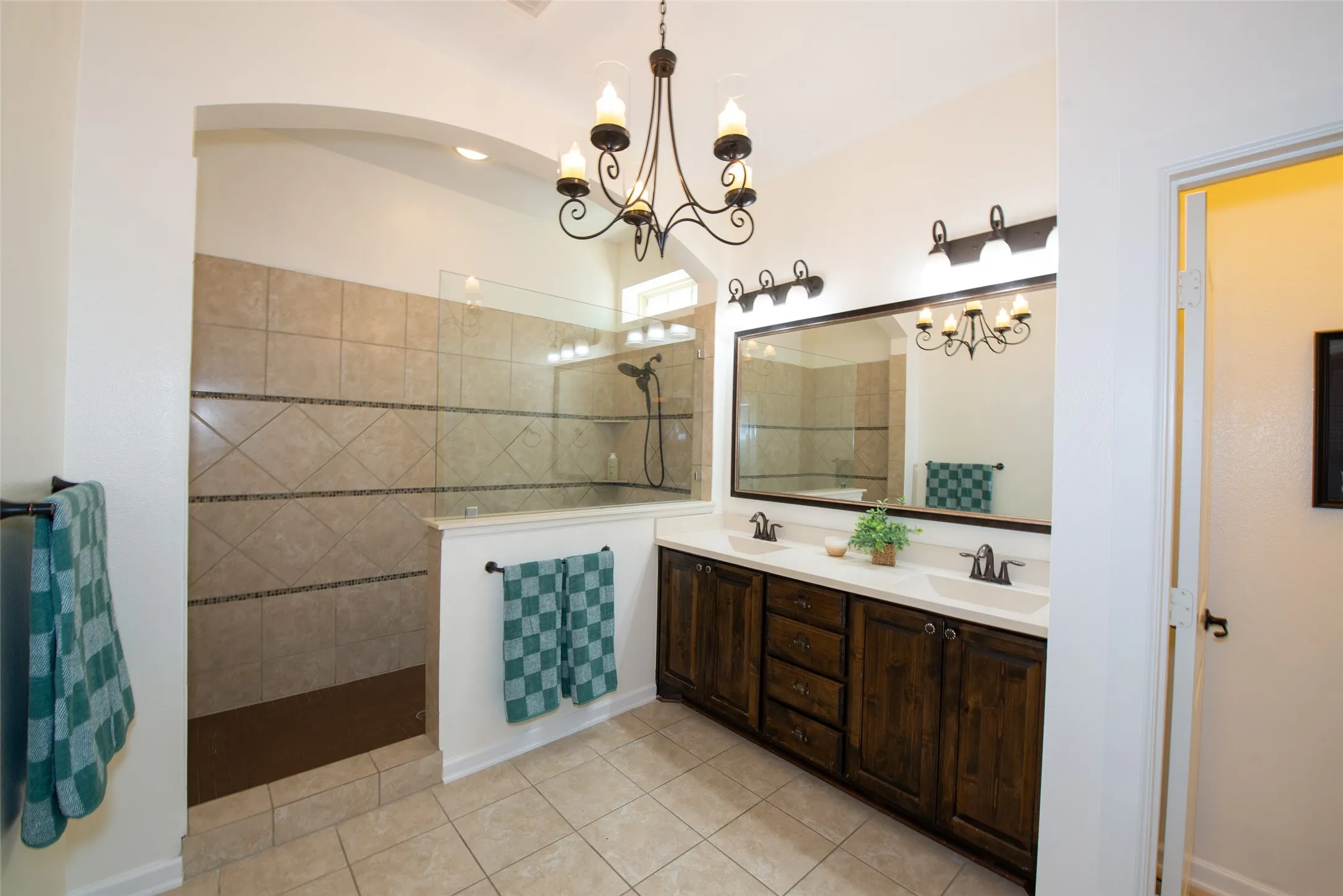 Spacious bathroom featuring a dual vanity with dark wood cabinetry and white countertops, a large framed mirror, and a walk-in shower with tiled walls