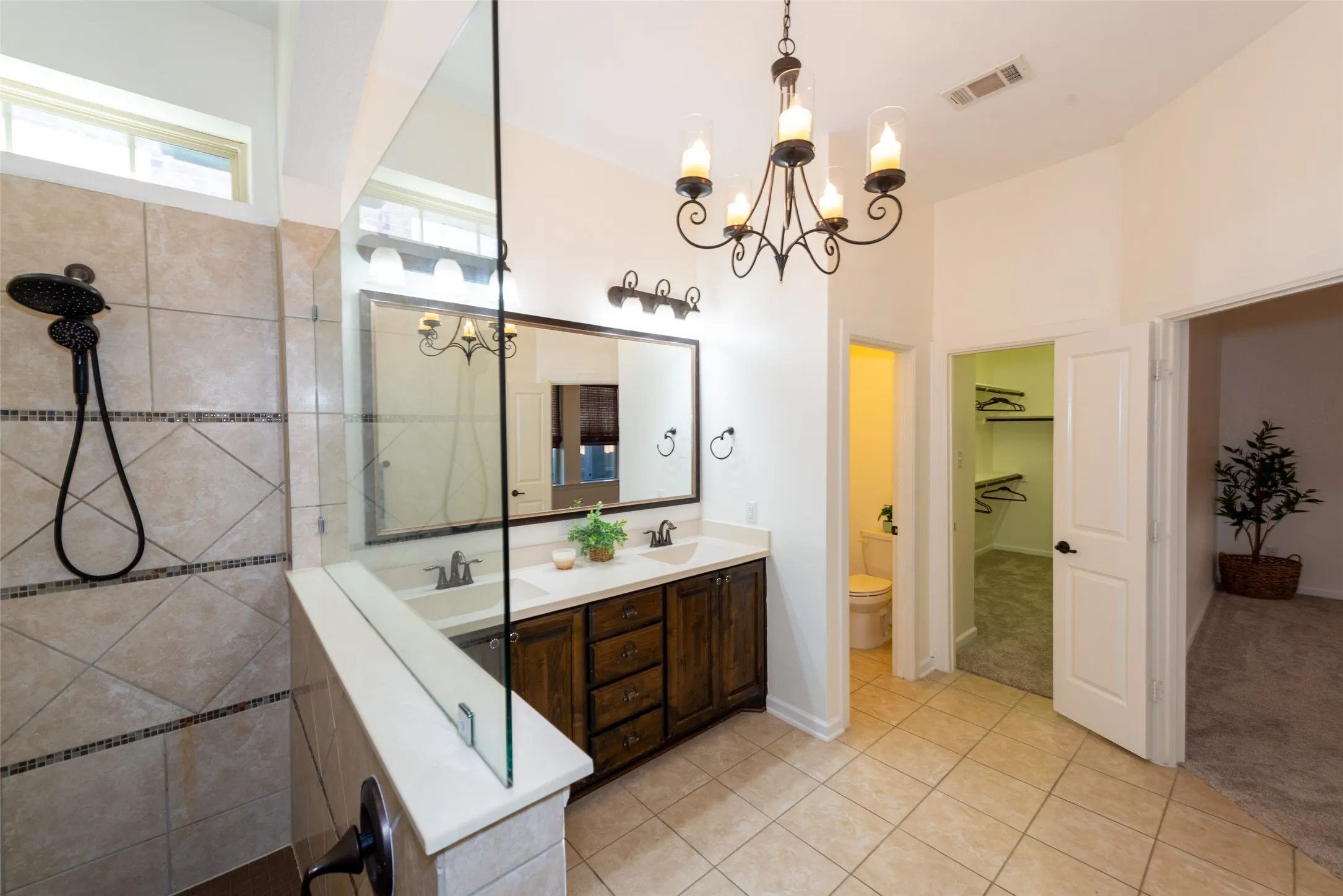 Spacious bathroom featuring a glass-enclosed shower with tile surround, double vanity with dark wood cabinetry and white countertops, tile flooring, and an ornamental chandelier