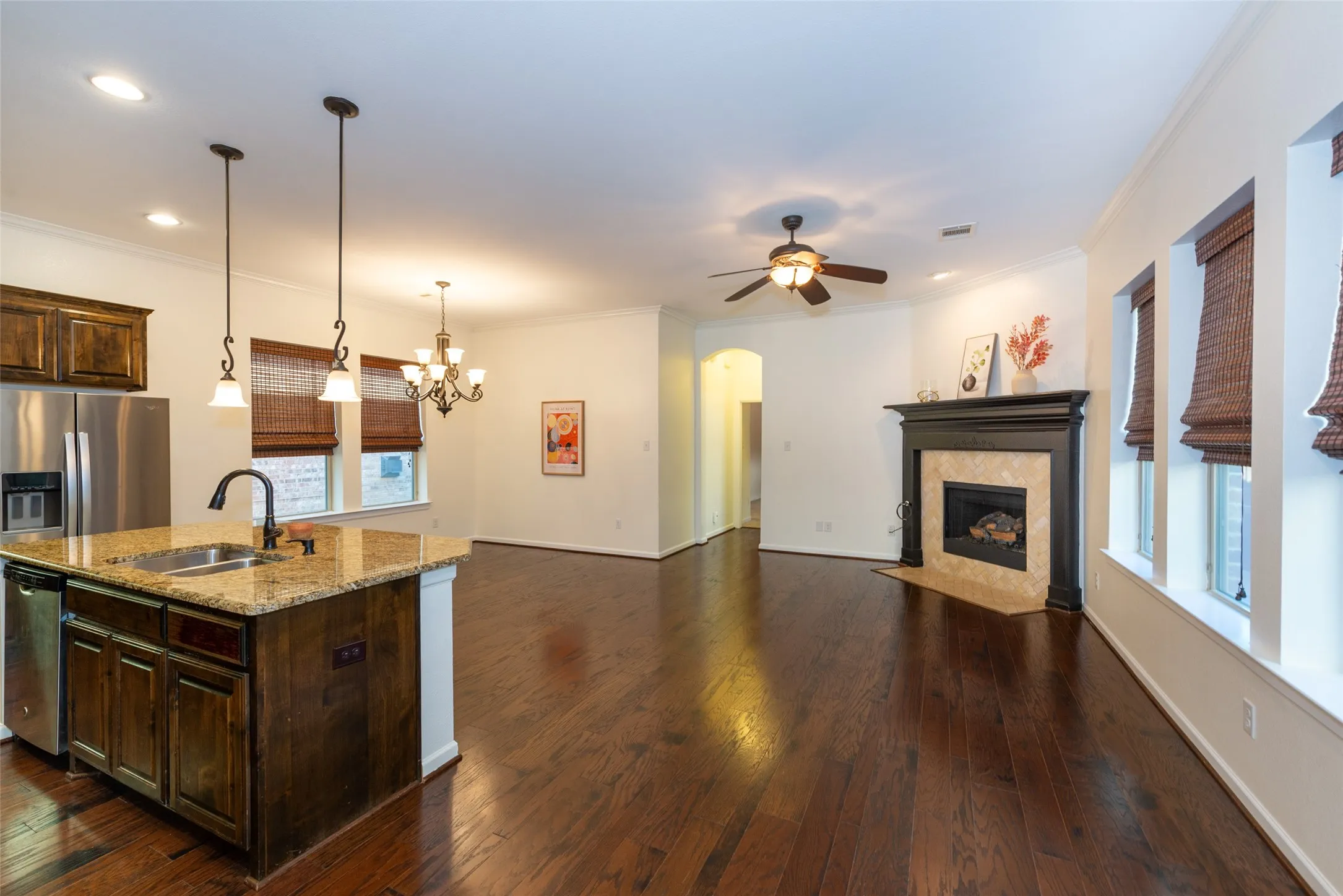 Open-concept living area featuring wood-finish flooring, a kitchen island with stone countertops, a stainless steel refrigerator, a fireplace with a dark wood mantel and stone surround, and multiple windows with woven blinds