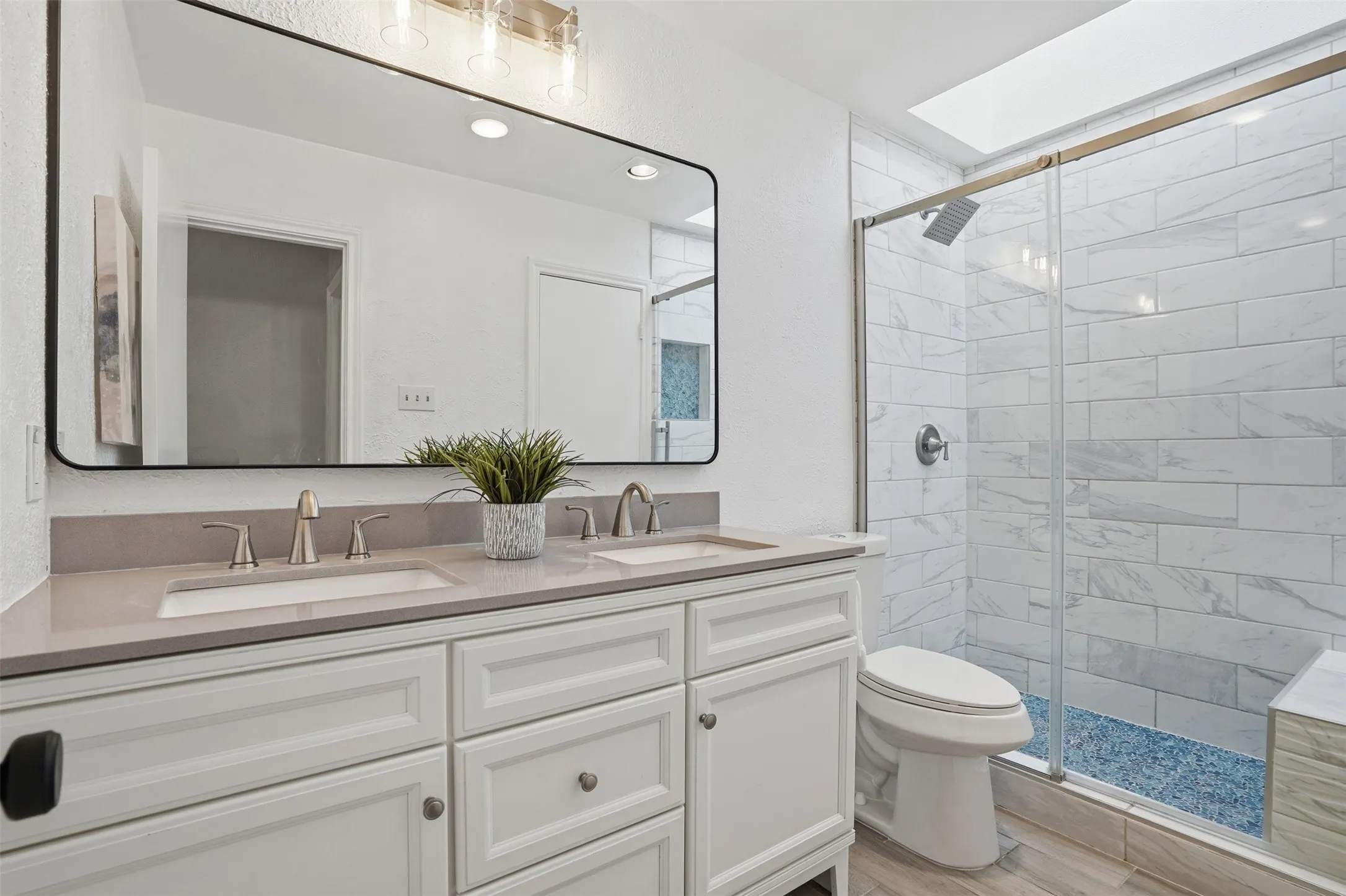 Bathroom featuring a dual-sink vanity with a light-colored countertop, an expansive frameless mirror, and a glass-enclosed shower with white tiled walls and a blue-tiled floor
