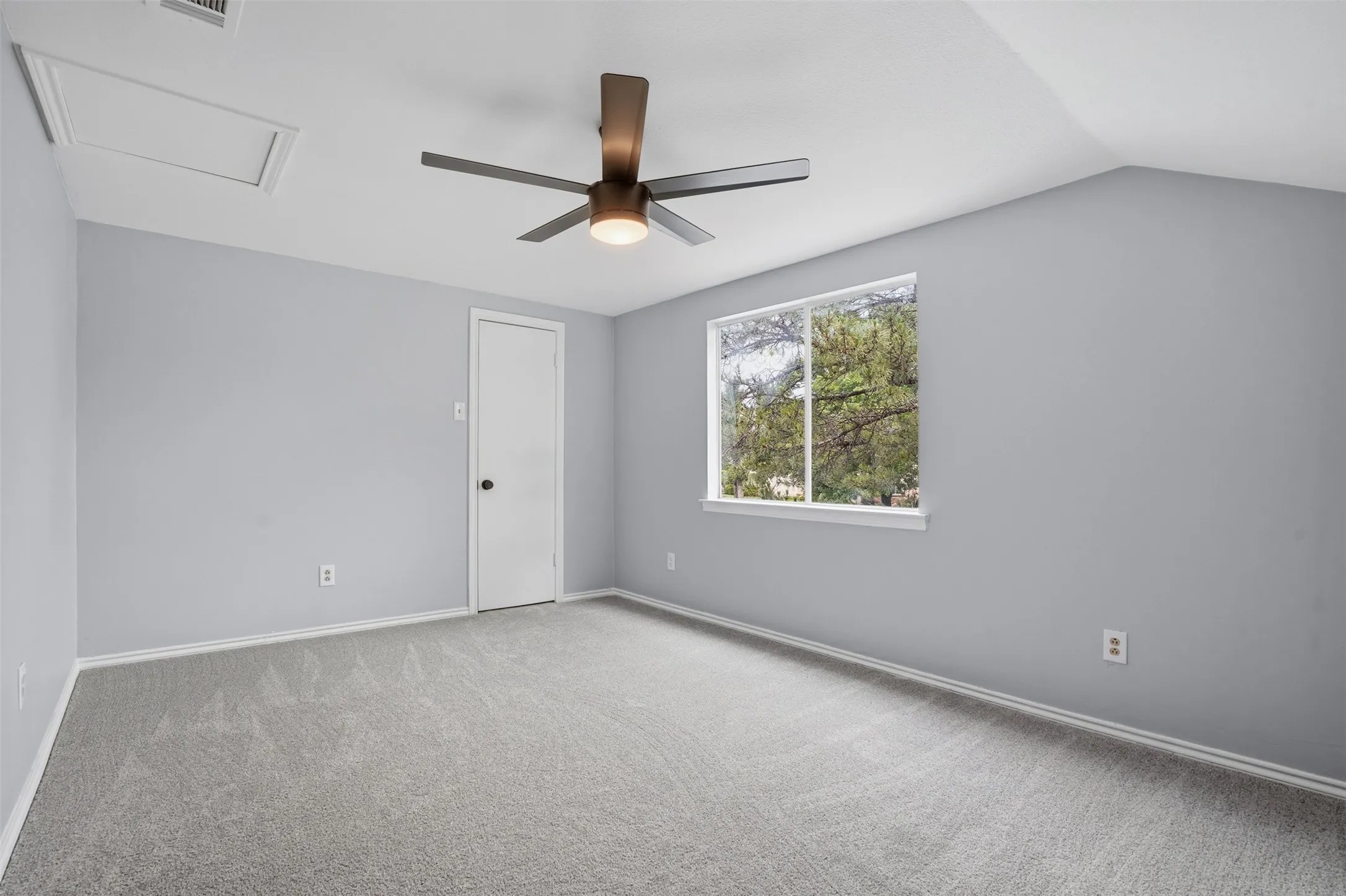 Carpeted room featuring a ceiling fan with integrated lighting, a window with exterior tree scenery, a white interior door with a black doorknob, and an attic access panel