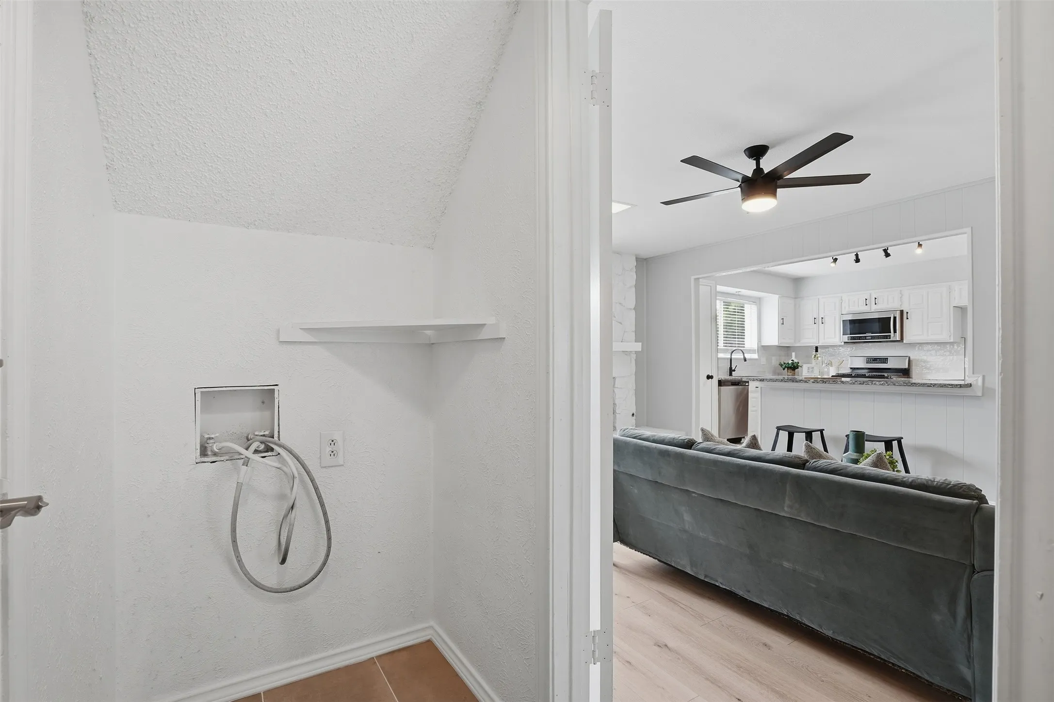 Functional laundry area with hookups and a corner shelf, offering a glimpse into the open-concept living space with a ceiling fan and kitchen beyond