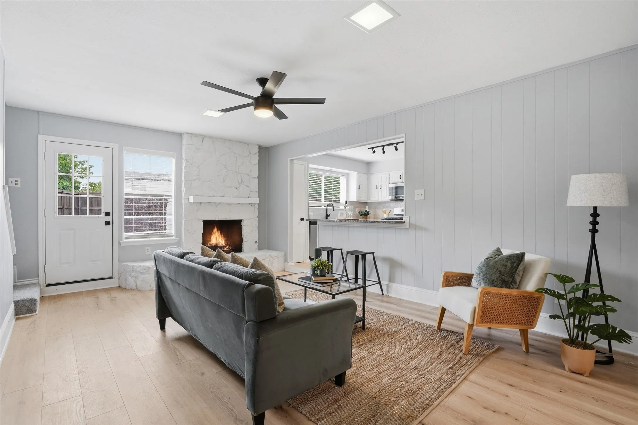 Open-concept living area featuring light wood-finish flooring, a white stone fireplace, and a ceiling fan with integrated lighting