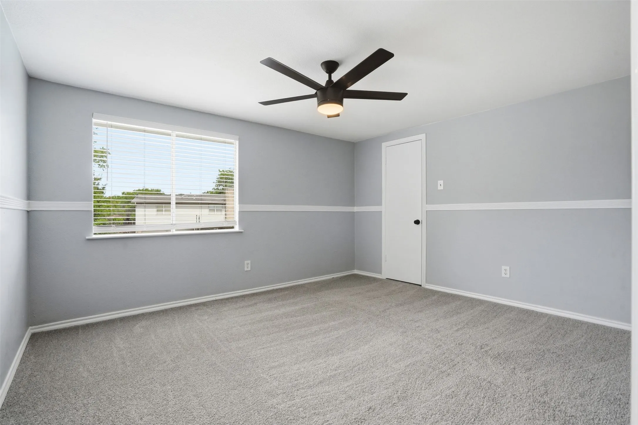 Carpeted interior room featuring a contemporary ceiling fan with integrated lighting, a large window with horizontal blinds, and a white interior door with dark hardware