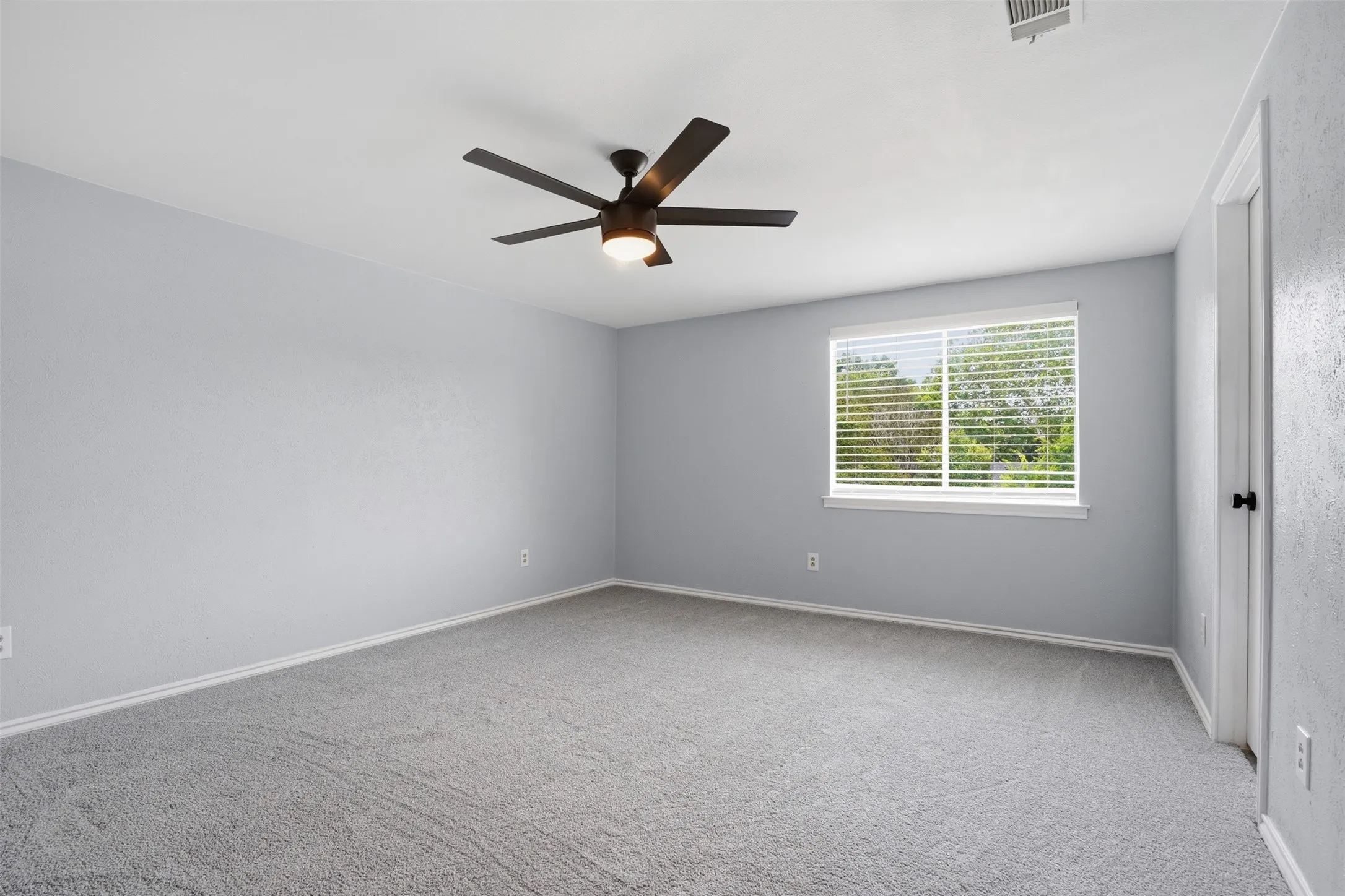 Spacious room featuring light gray walls, gray carpet, and a window with blinds overlooking greenery