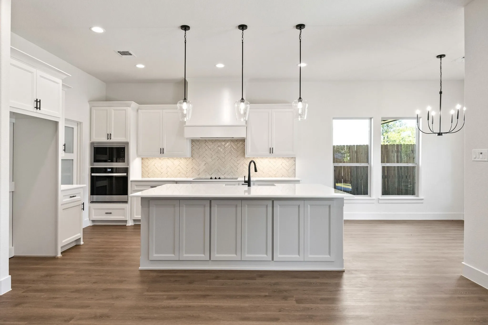 Open-concept kitchen featuring an oversized island with a built-in sink, white shaker cabinetry, herringbone tile backsplash, integrated double ovens, and wood-finish flooring
