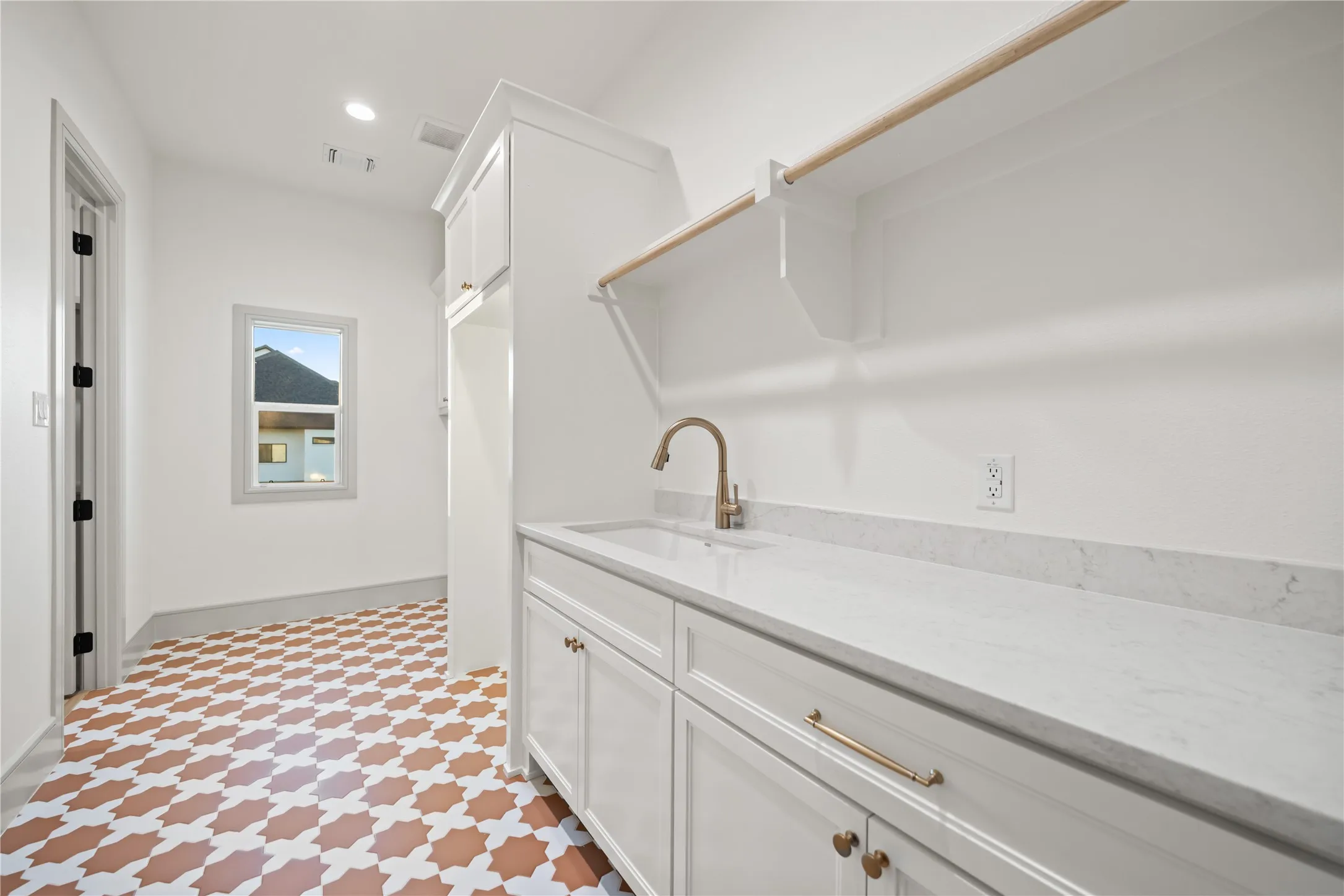 Laundry room featuring intricate patterned tile flooring, a white vanity with a rectangular undermount sink, a gold-finish faucet, white cabinetry with gold-finish hardware, and a built-in shelving unit with a hanging rod