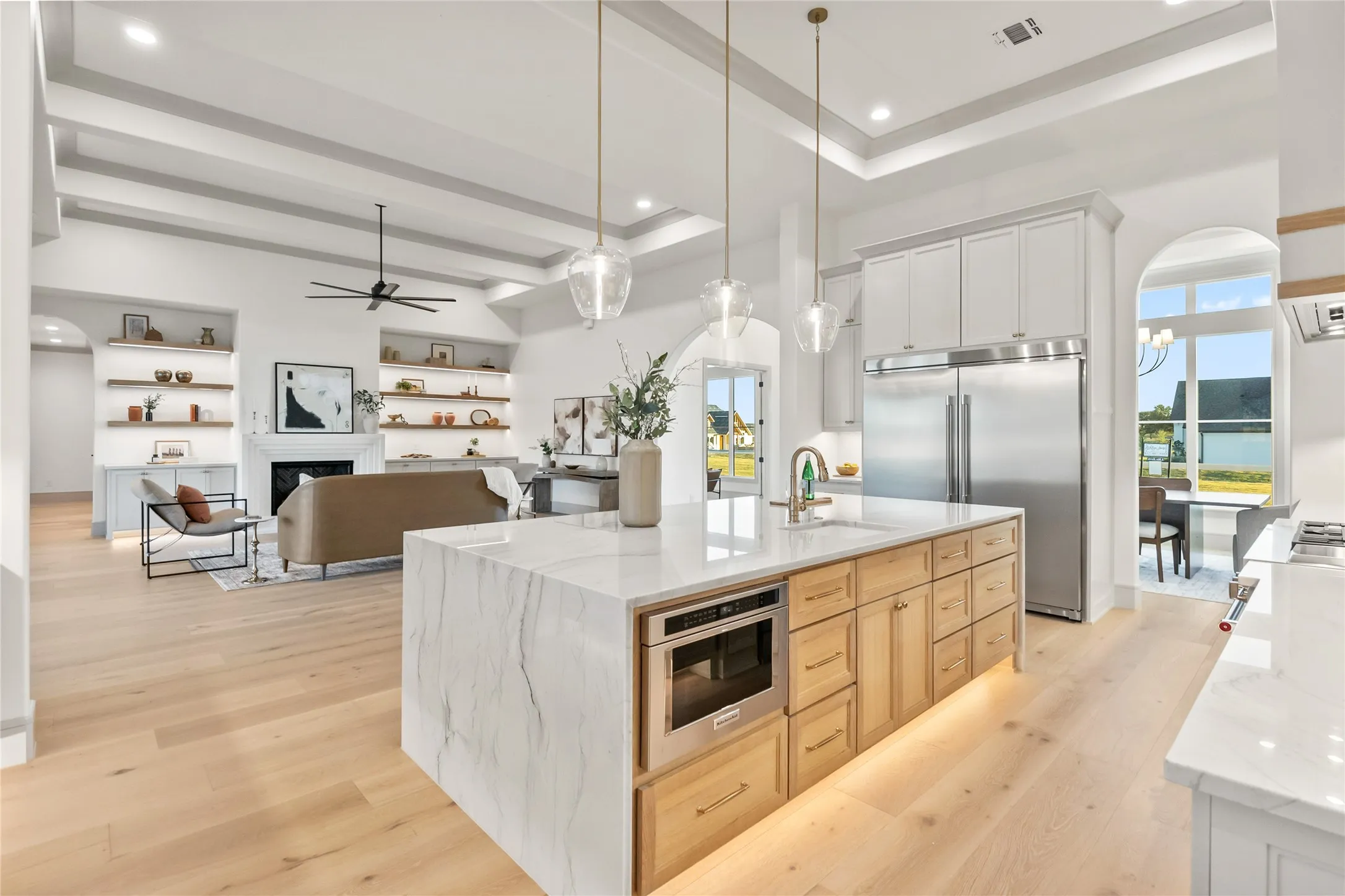 Open-concept kitchen and living area featuring light wood-finish flooring, a kitchen island with a waterfall countertop and integrated microwave, stainless steel appliances, and a coffered ceiling
