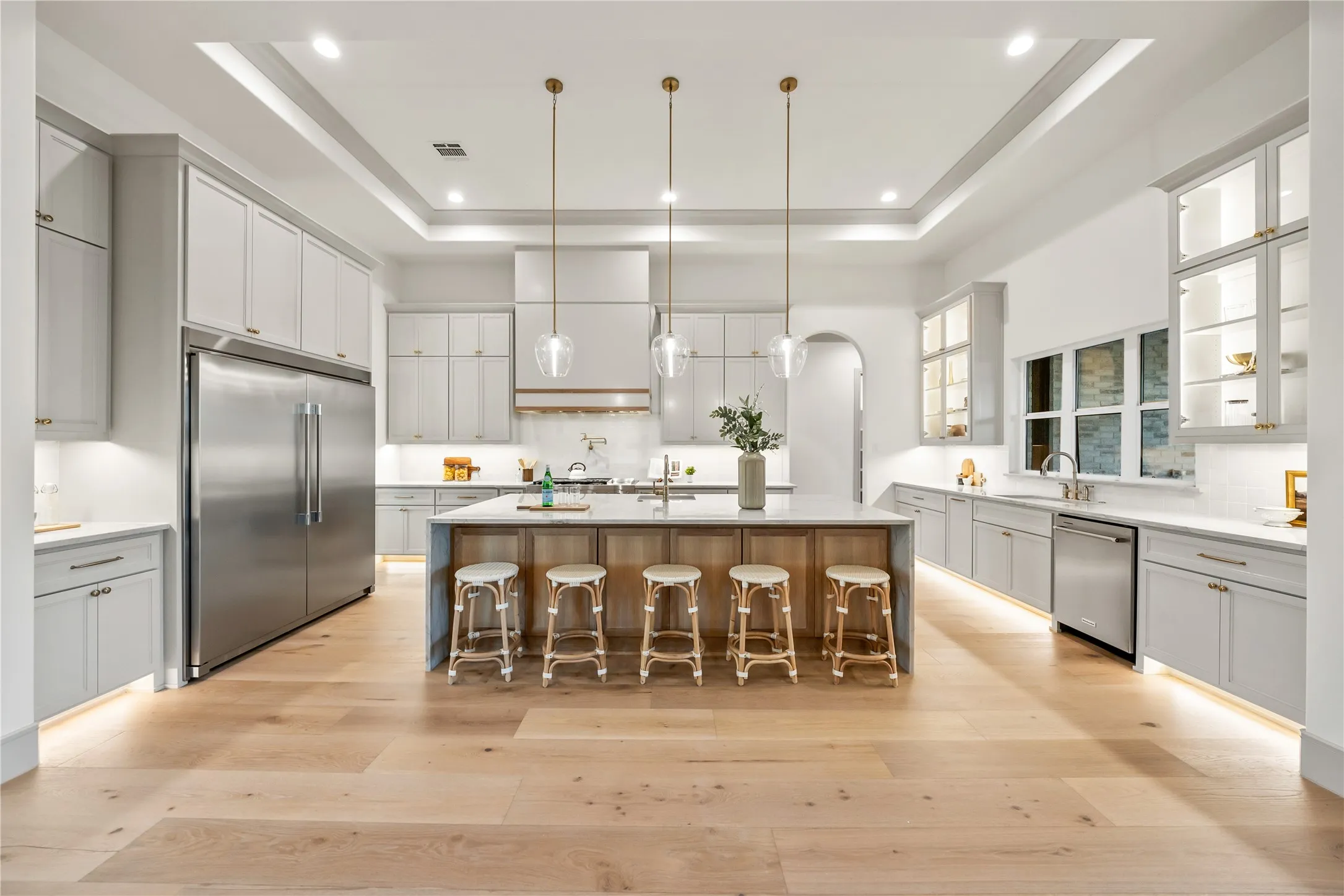 Spacious kitchen featuring light wood-finish flooring, a large center island with seating, extensive grey cabinetry, stainless steel appliances, and tray ceilings with recessed lighting