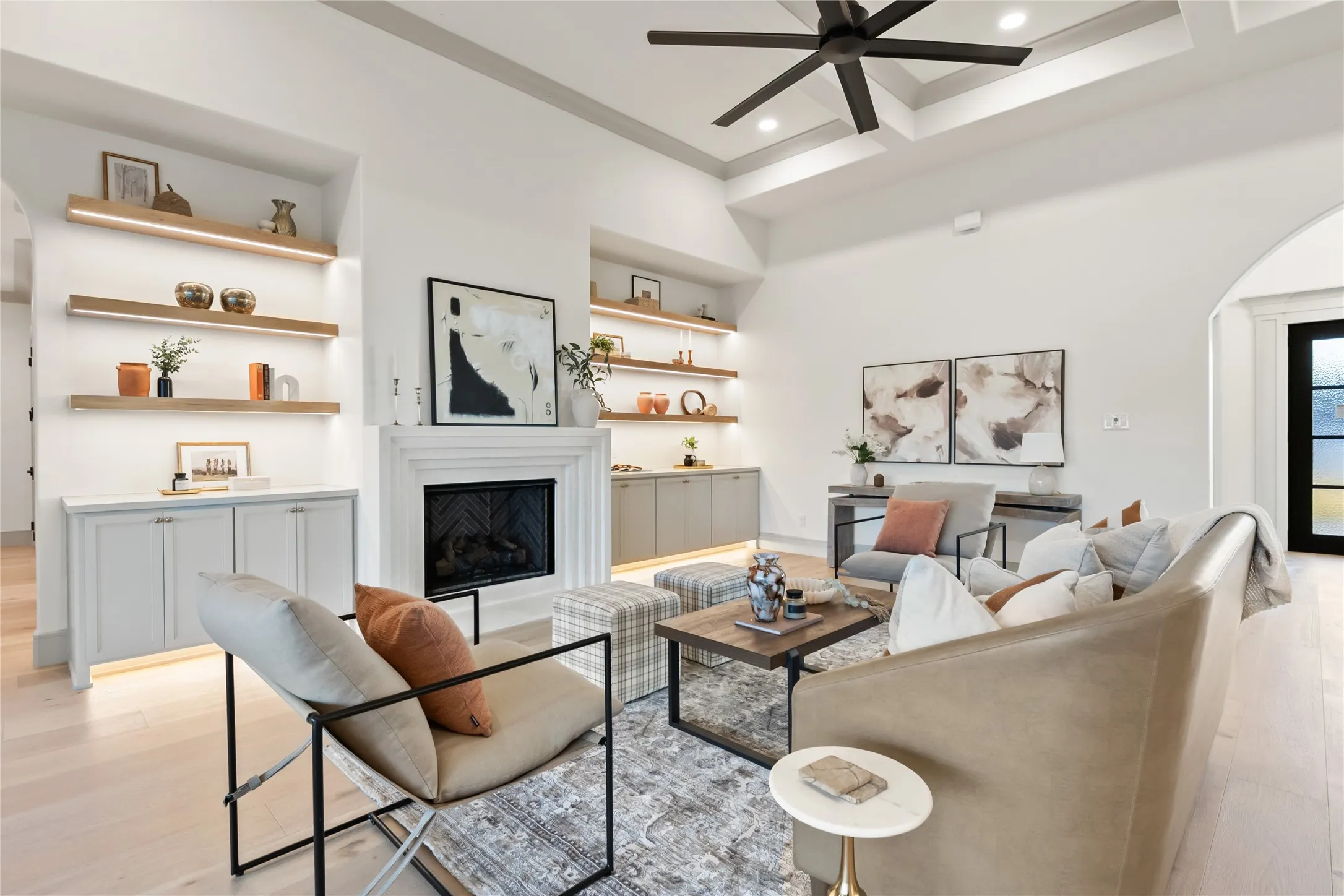 Spacious living area featuring a fireplace with a detailed mantel, built-in cabinetry with under-cabinet lighting, tray ceiling with a ceiling fan, and light wood-finish flooring