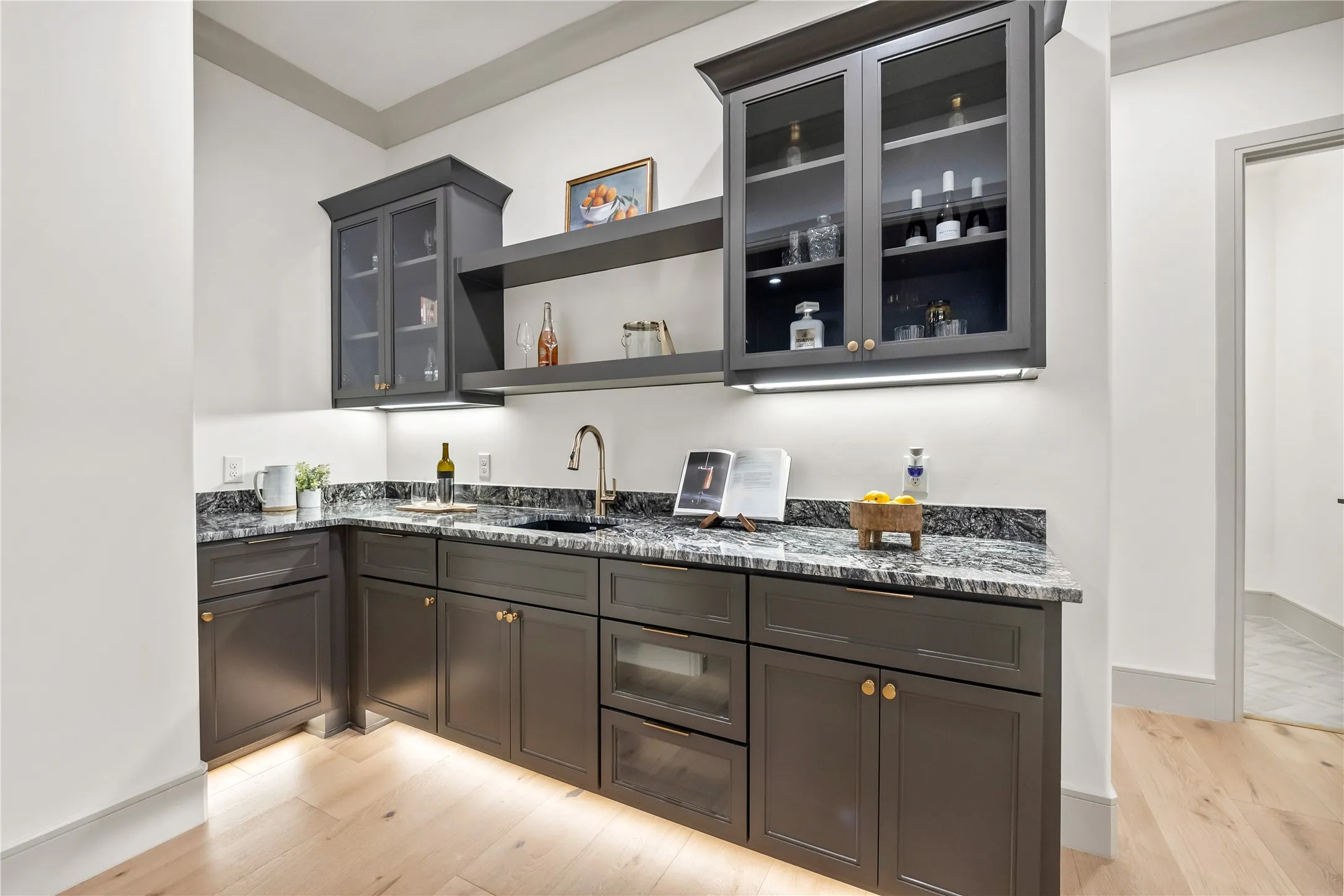 Built-in wet bar featuring dark cabinetry, granite countertops, an undermount sink with a gooseneck faucet, display shelving, and light wood-finish flooring