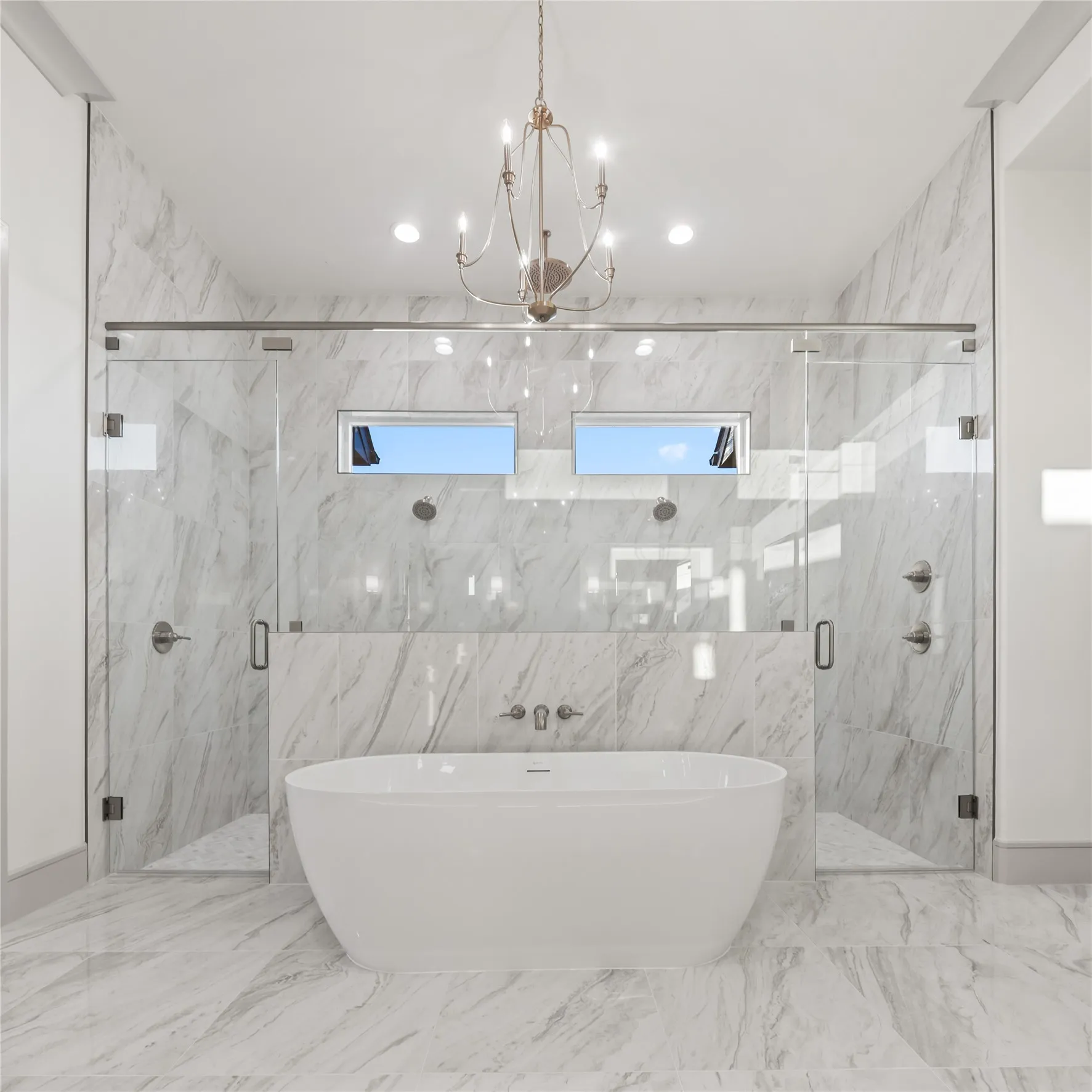 Freestanding soaking tub centered in a spacious bathroom featuring floor-to-ceiling marbled tile, dual glass-enclosed showers, and a chandelier