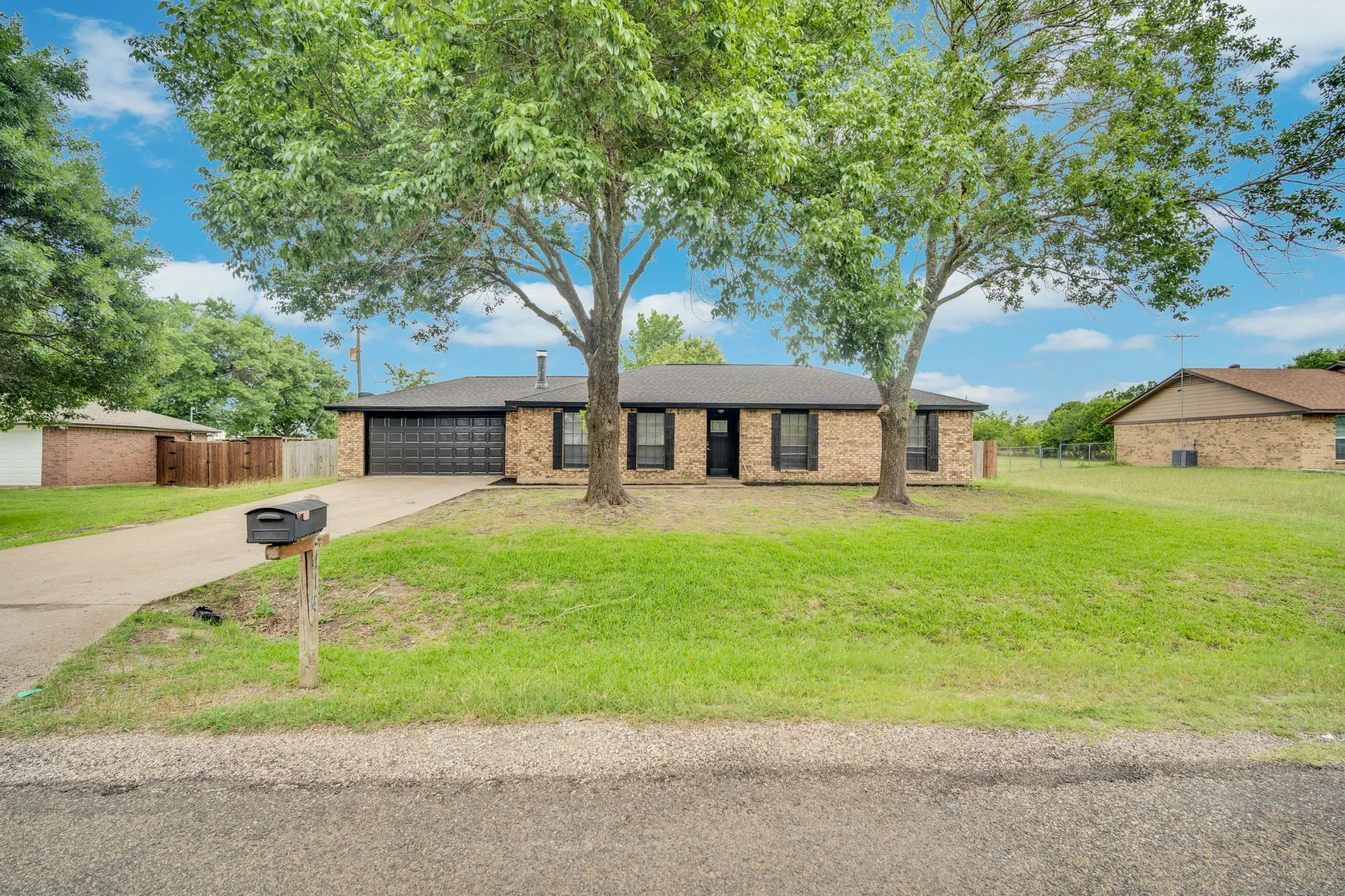 Brick exterior home featuring a two-car garage, mature front yard trees, a concrete driveway, and a mailbox