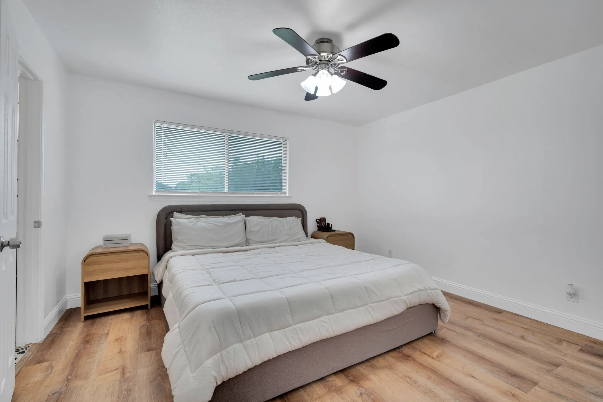 Bedroom featuring wood-finish flooring, a contemporary ceiling fan with integrated lighting, a window with horizontal blinds, and a white interior door with a brushed nickel knob
