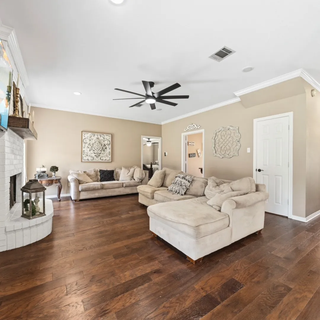 Spacious living area featuring rich wood-finish flooring, a white brick fireplace with a wood mantle, crown molding, and recessed lighting