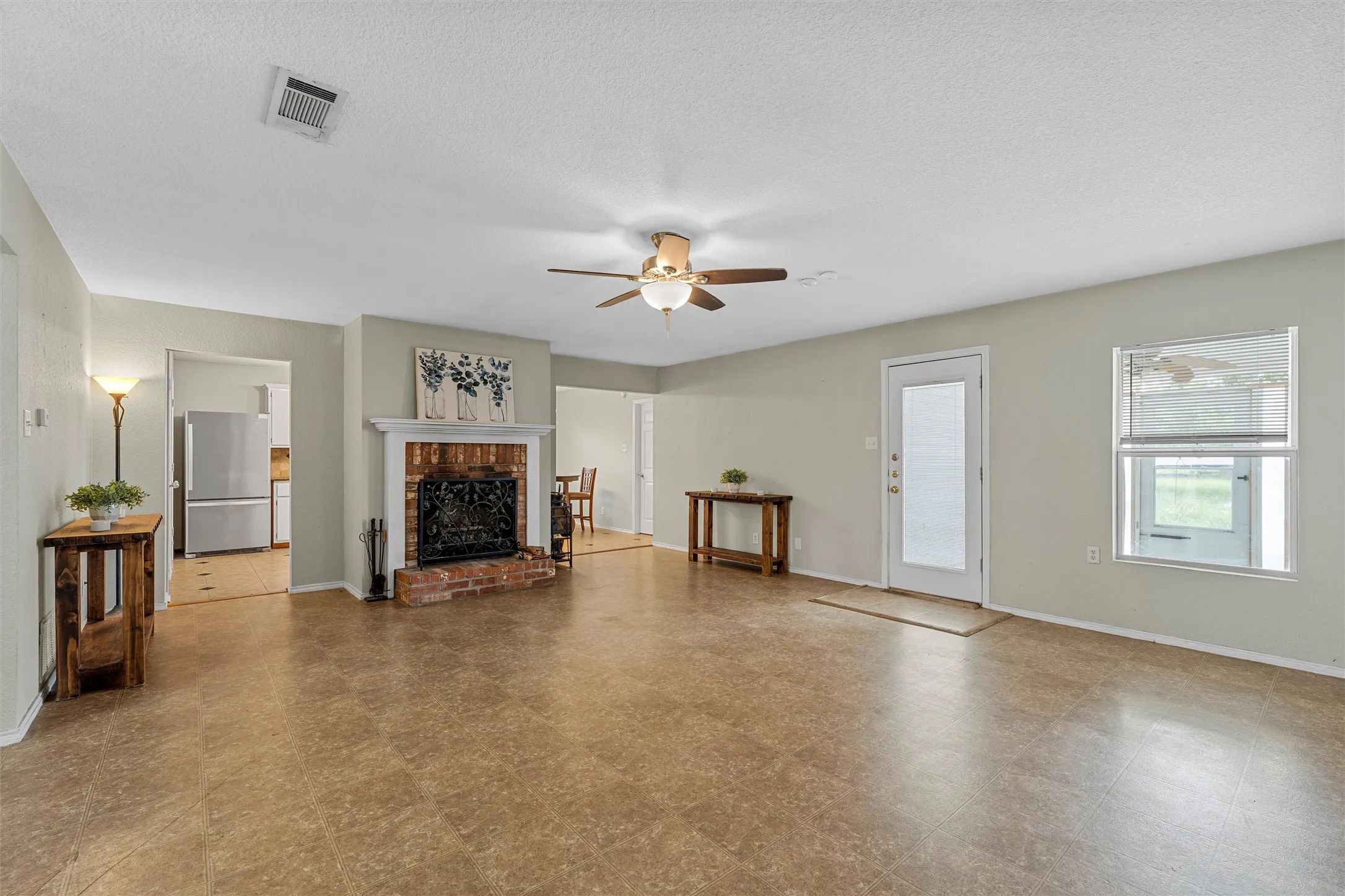 Spacious living area featuring a brick fireplace with a white mantel, ceiling fan with light fixture, and warm-toned flooring