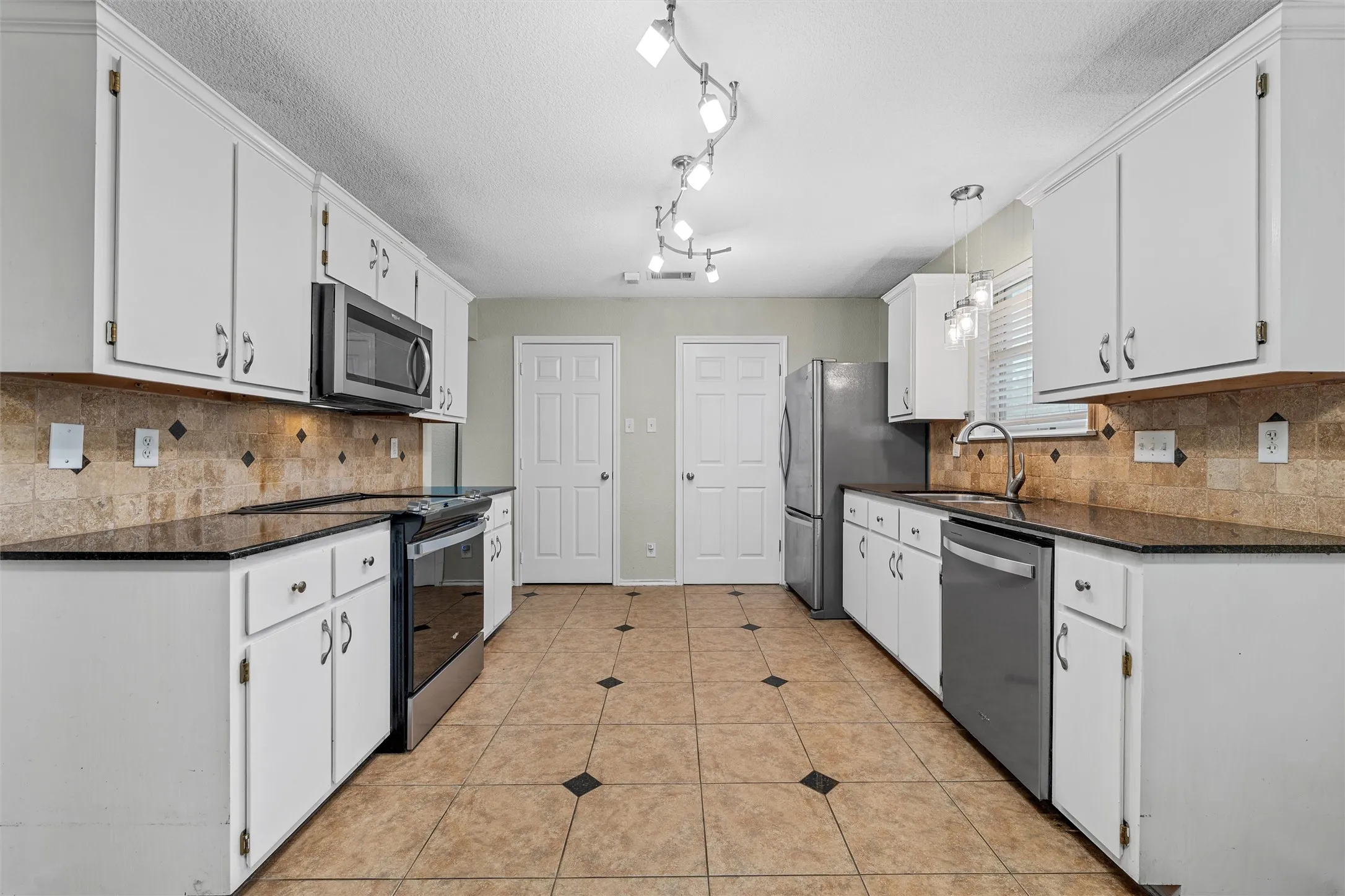 Galley kitchen featuring white cabinetry, dark countertops, stainless steel appliances, a tiled backsplash with diamond accents, and ceramic tile flooring