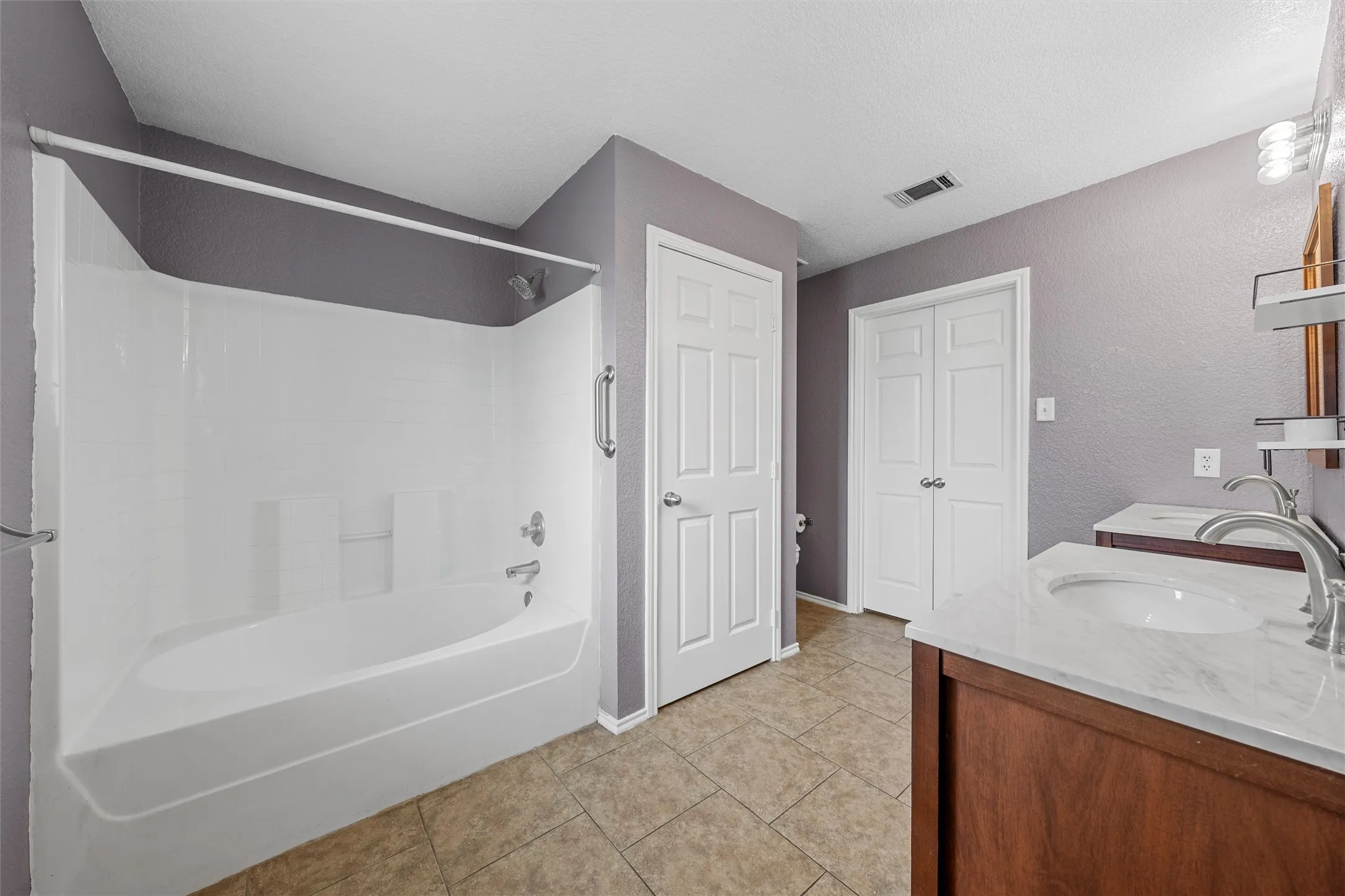 Bathroom featuring a white tub/shower combination, dual vanities with white countertops, wood-finish cabinetry, and tiled flooring