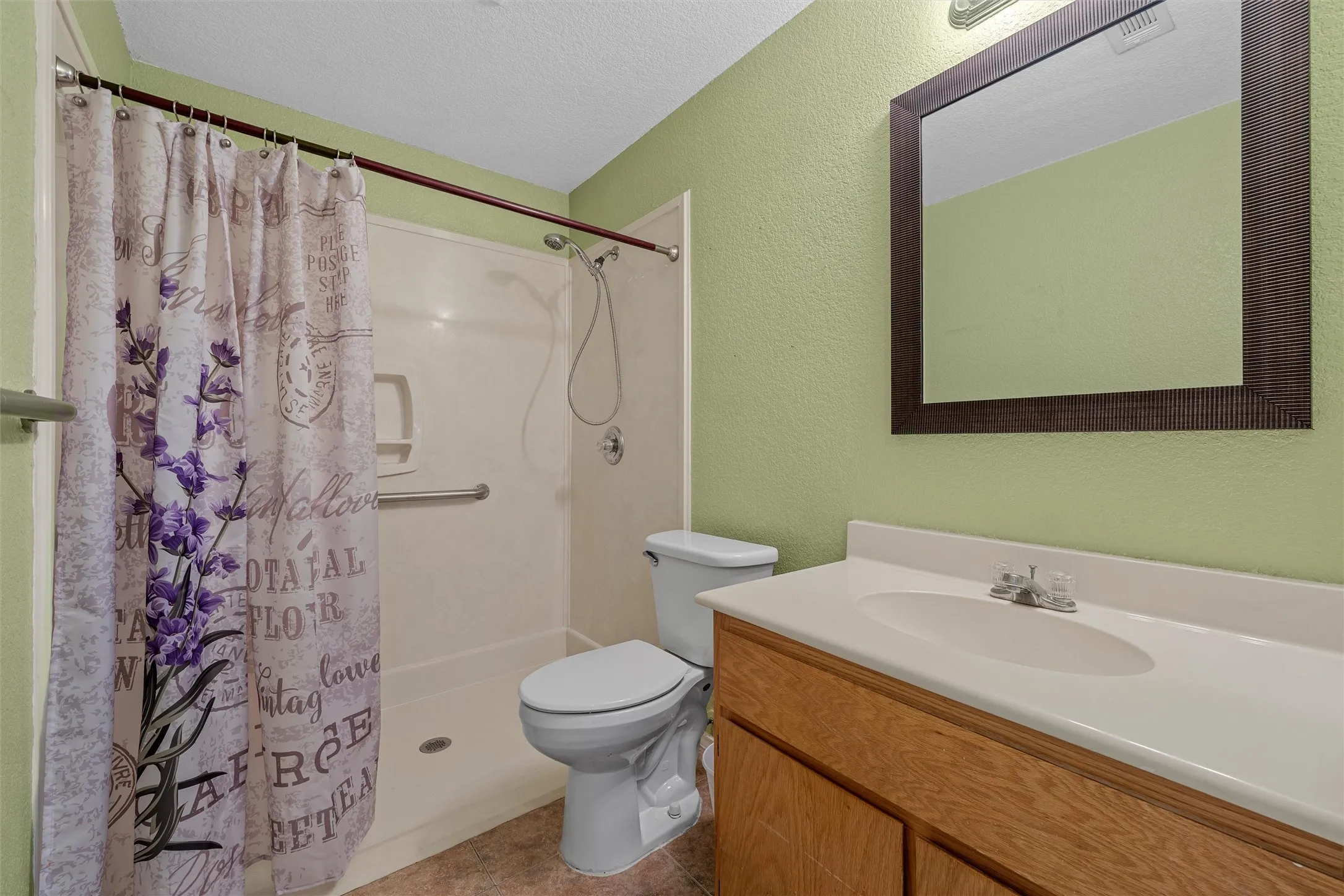 Bathroom featuring a walk-in shower with grab bars, a single-sink vanity with wood-finish cabinetry, a white countertop, and a framed mirror