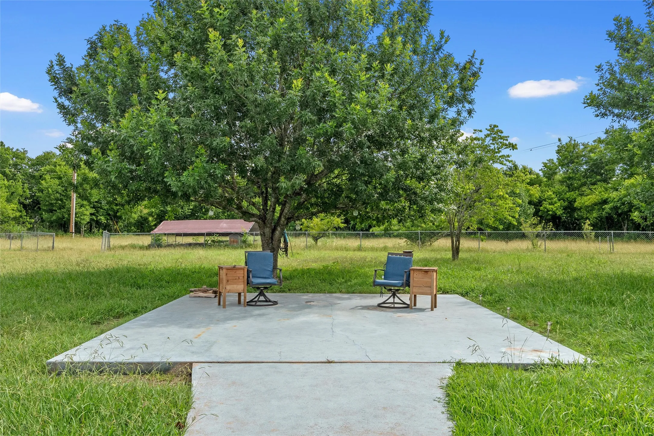 Concrete patio area featuring a mature shade tree, expansive grassy grounds, and perimeter fencing