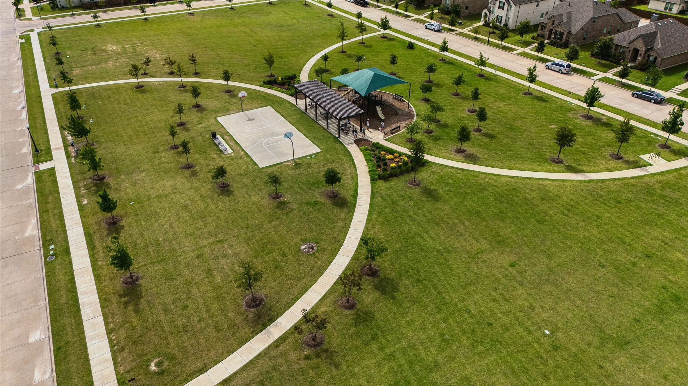 Expansive community green space featuring a basketball court, covered playground structure with green shade canopy, and paved walking paths