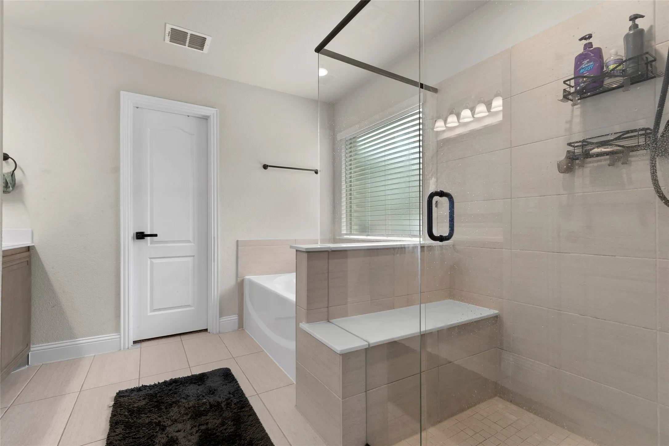Glass-enclosed shower with a built-in bench, complemented by light-toned wall tiling and a window with blinds